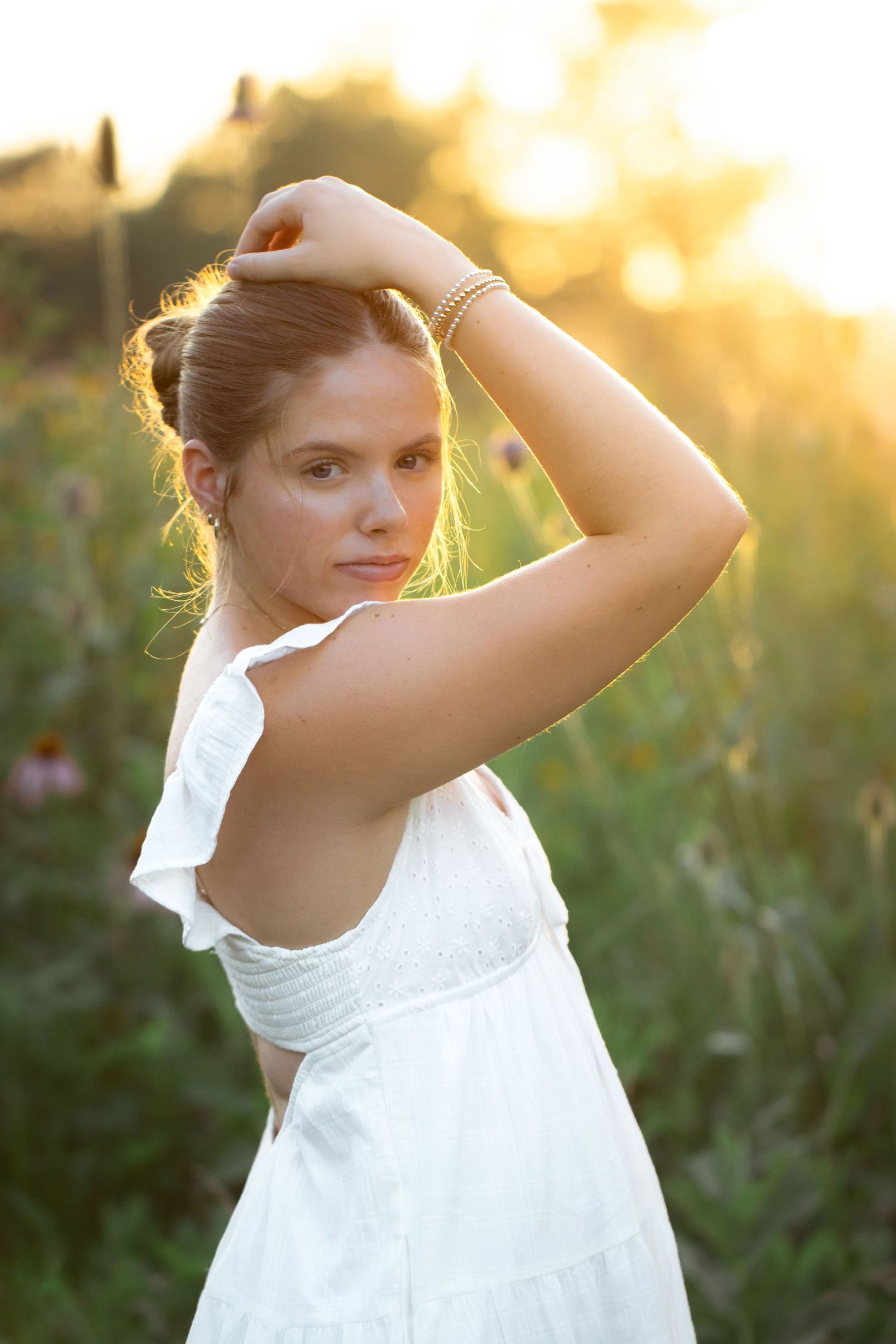 Young woman in white dress poses in golden hour sunlight among wildflowers, hand raised gracefully above her head.