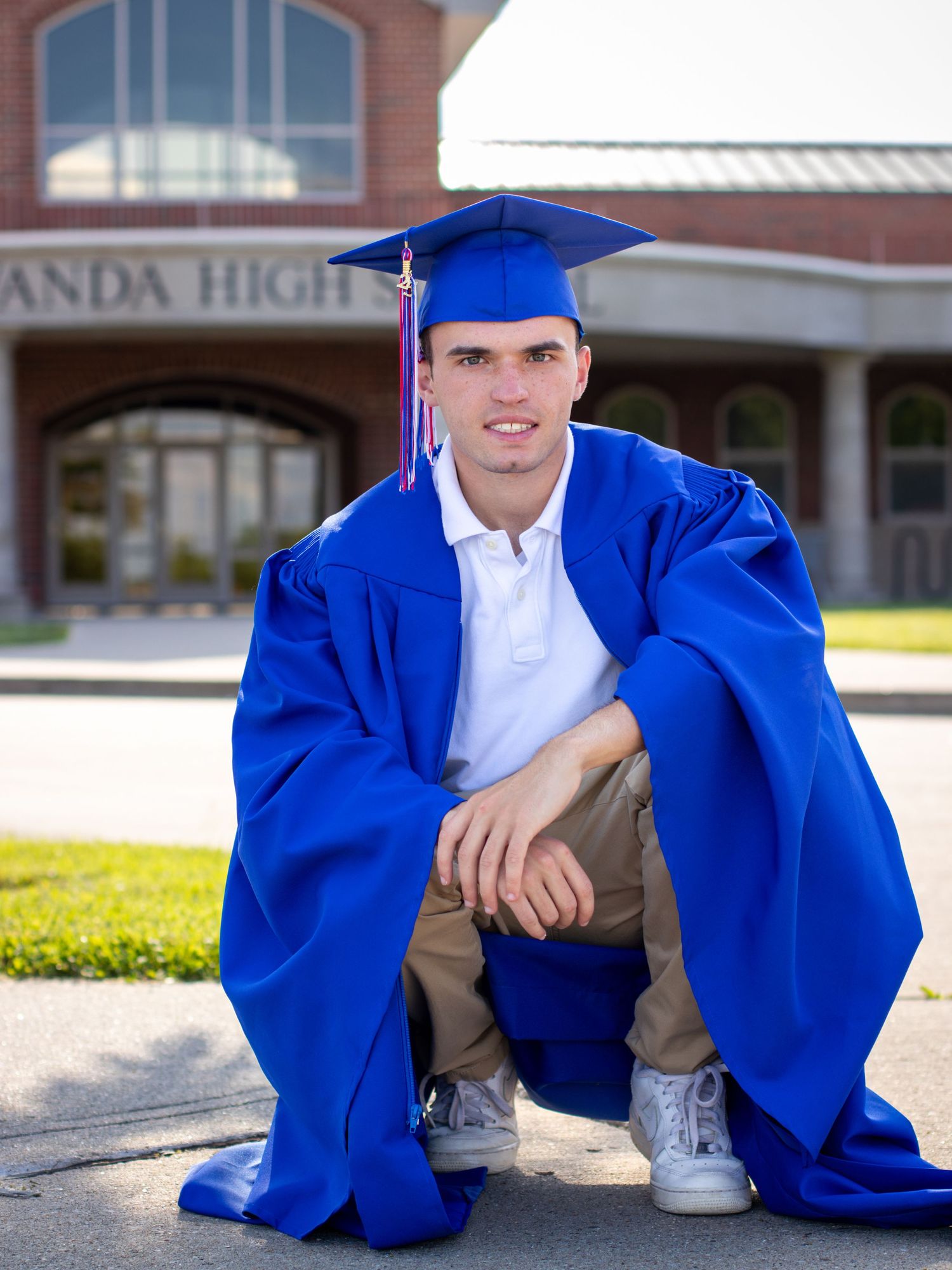 Graduate in blue cap and gown sitting casually outdoors near school building.