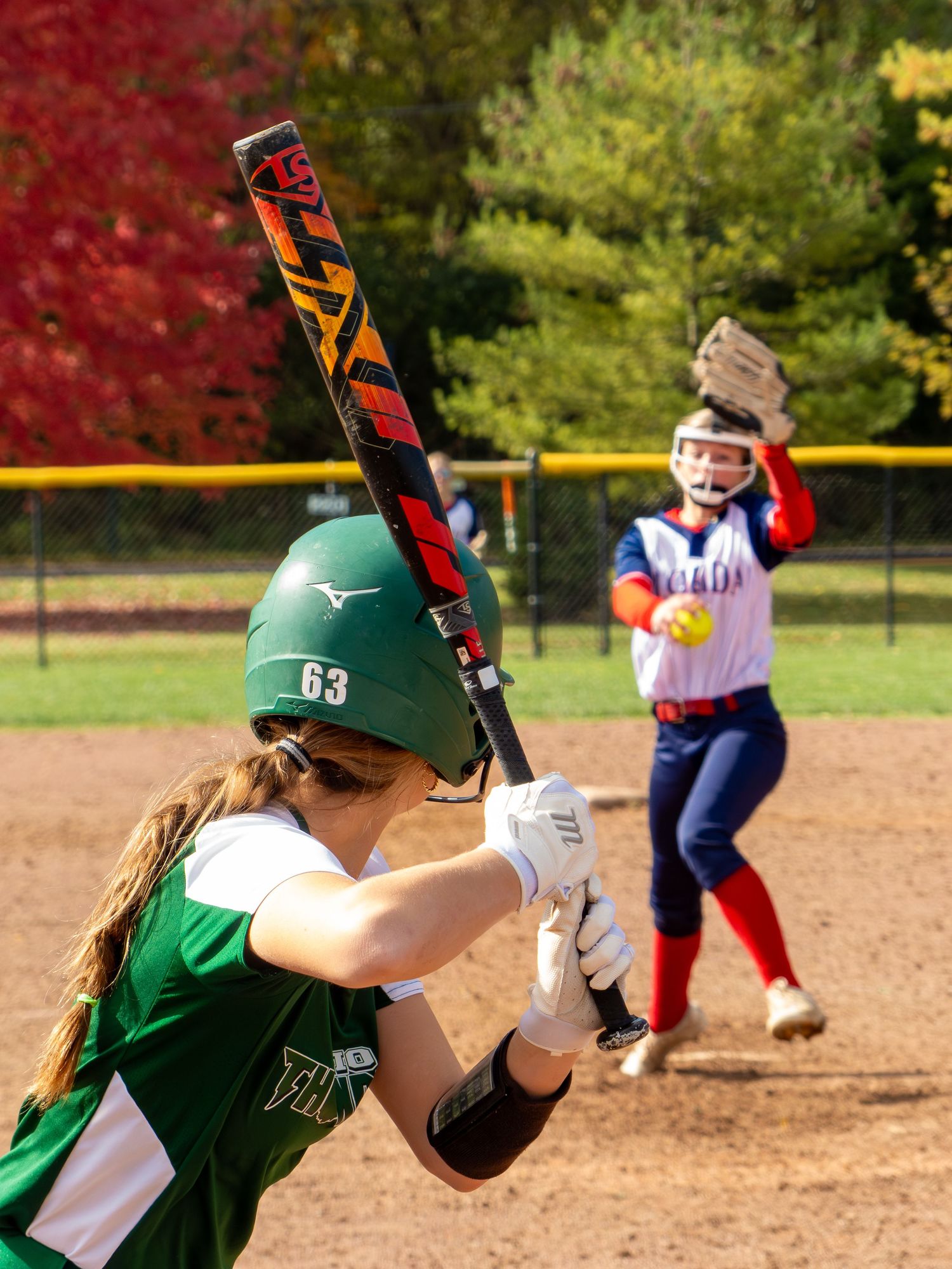 A softball player in green uniform takes a batting stance at home plate during a game on a sunny autumn day.