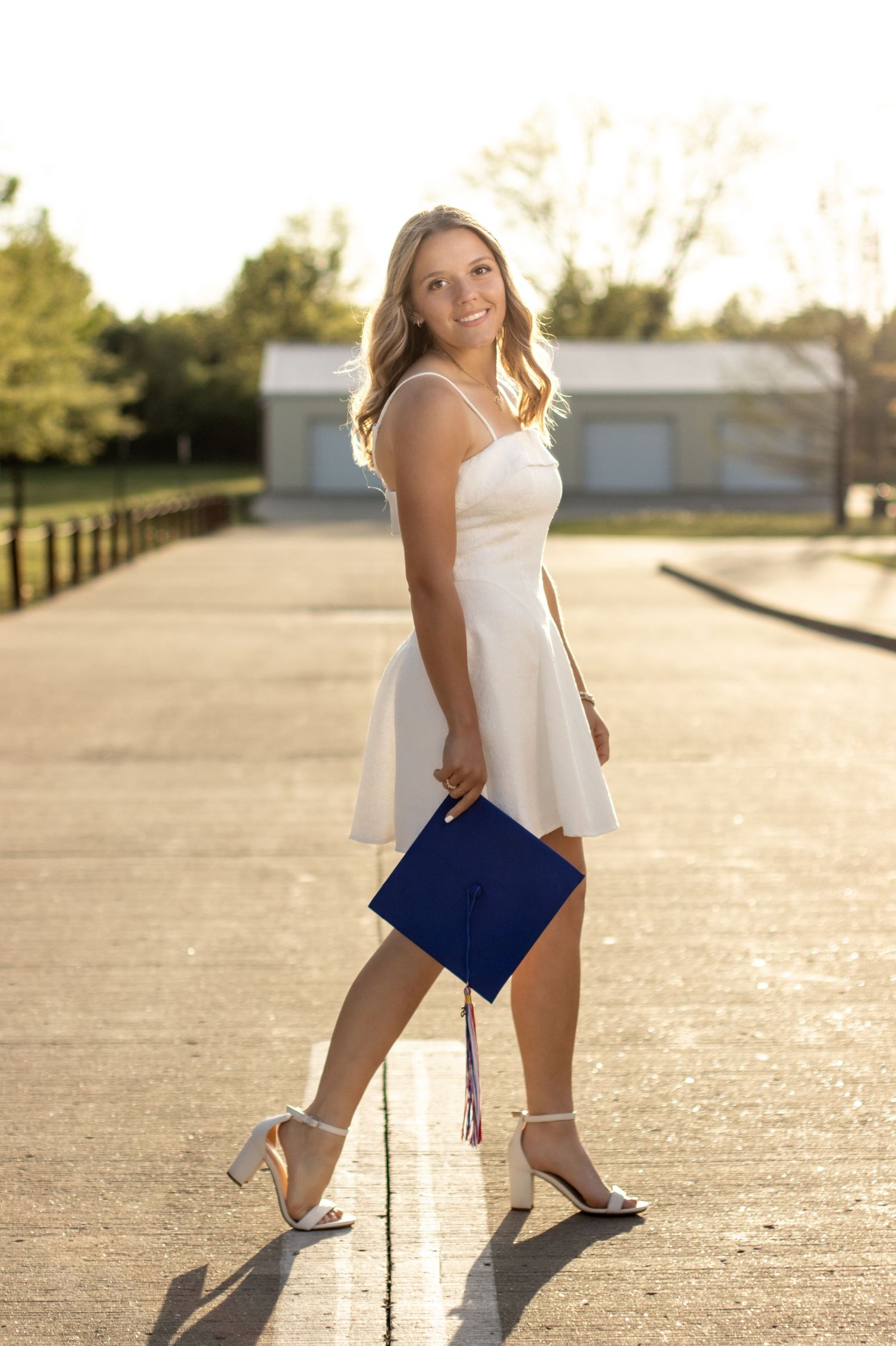 Young woman in white dress holding blue graduation cap poses outdoors in golden hour sunlight.