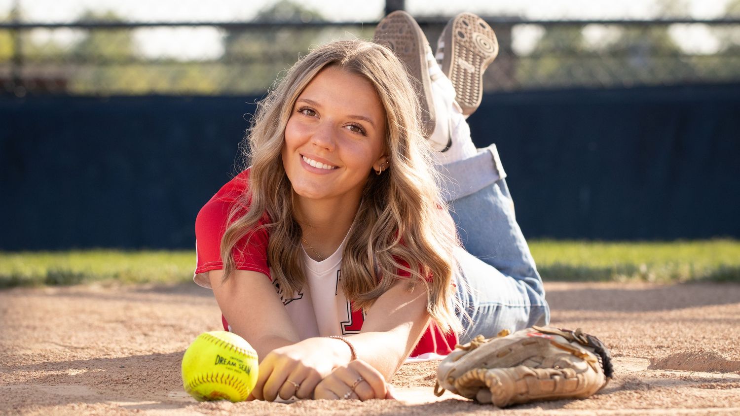 Young woman lying on a baseball field smiling, with a softball and glove beside her.