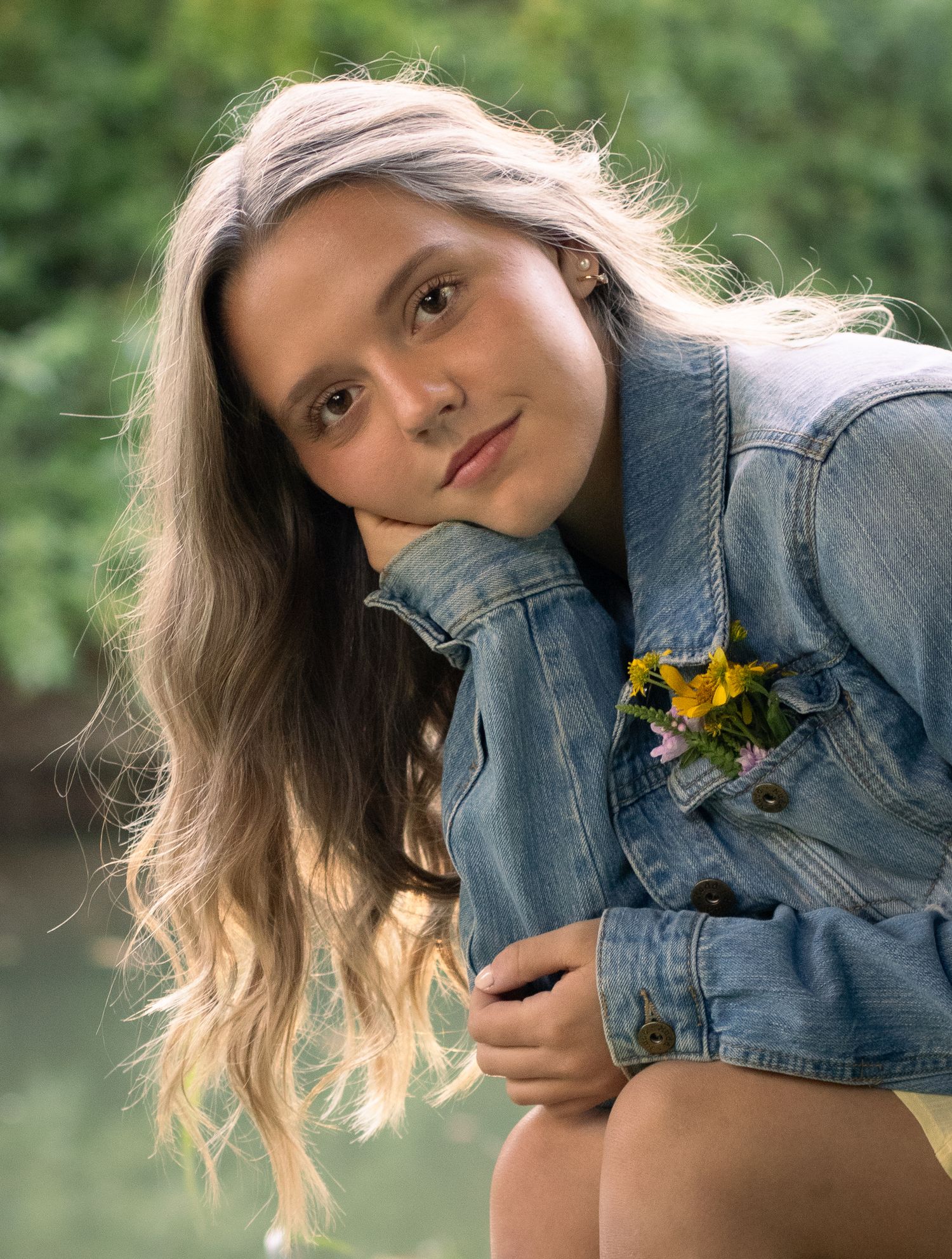 Young woman with long blonde hair wearing a denim jacket with yellow flowers, posing outdoors.