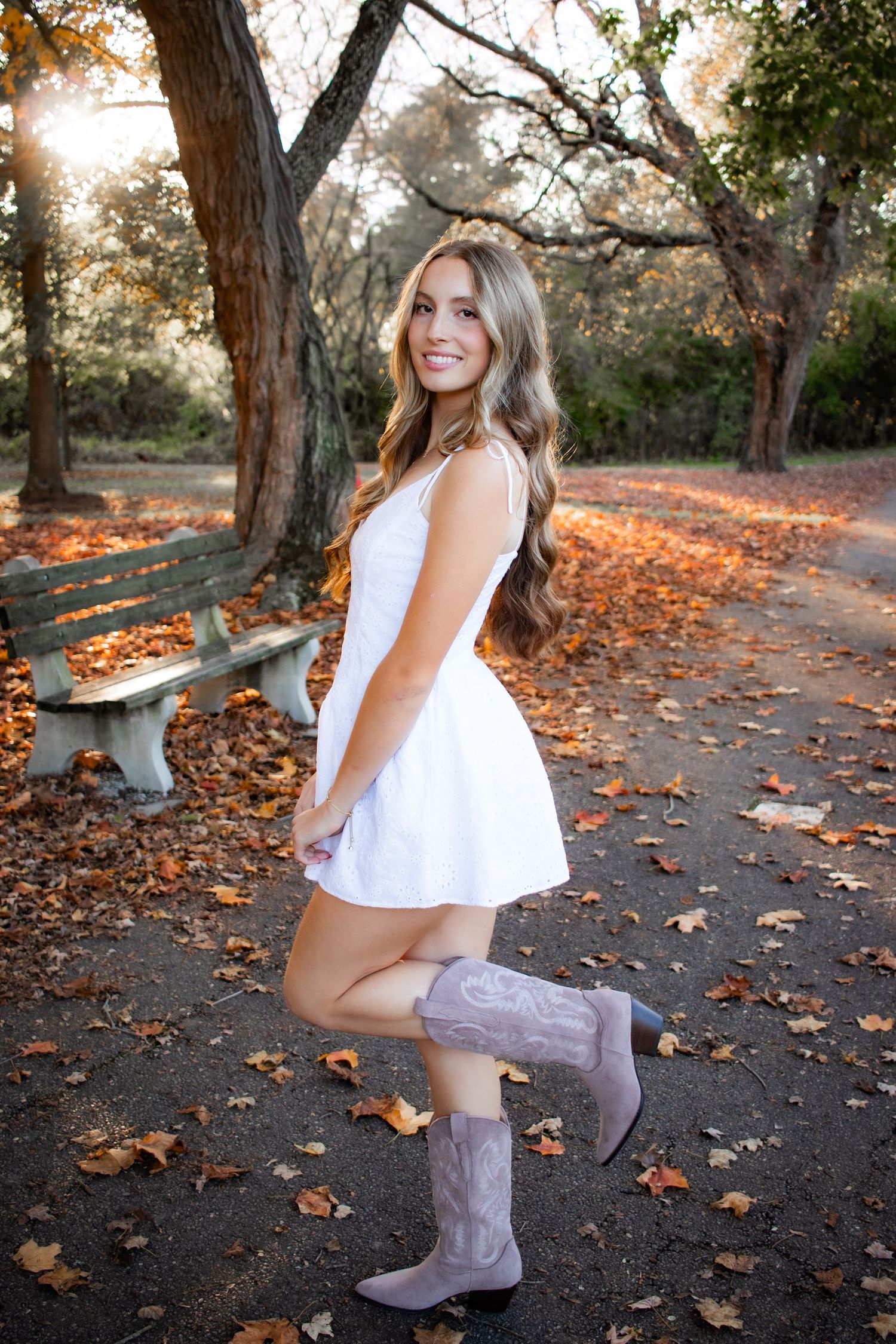 Person in white dress and boots poses on path covered with fallen leaves.