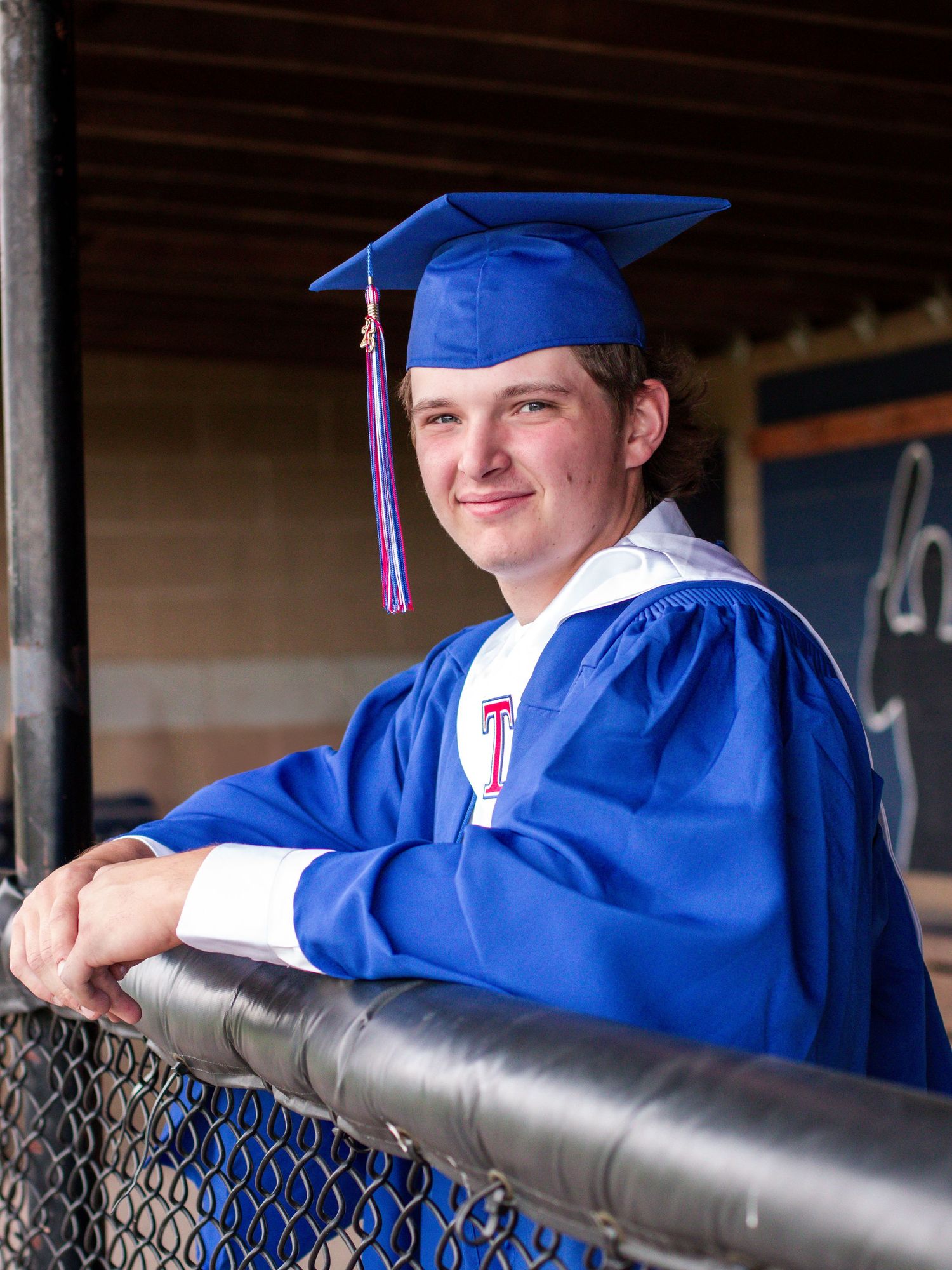 Young male graduate smiling in blue cap and gown, leaning on a fence in a dugout setting.