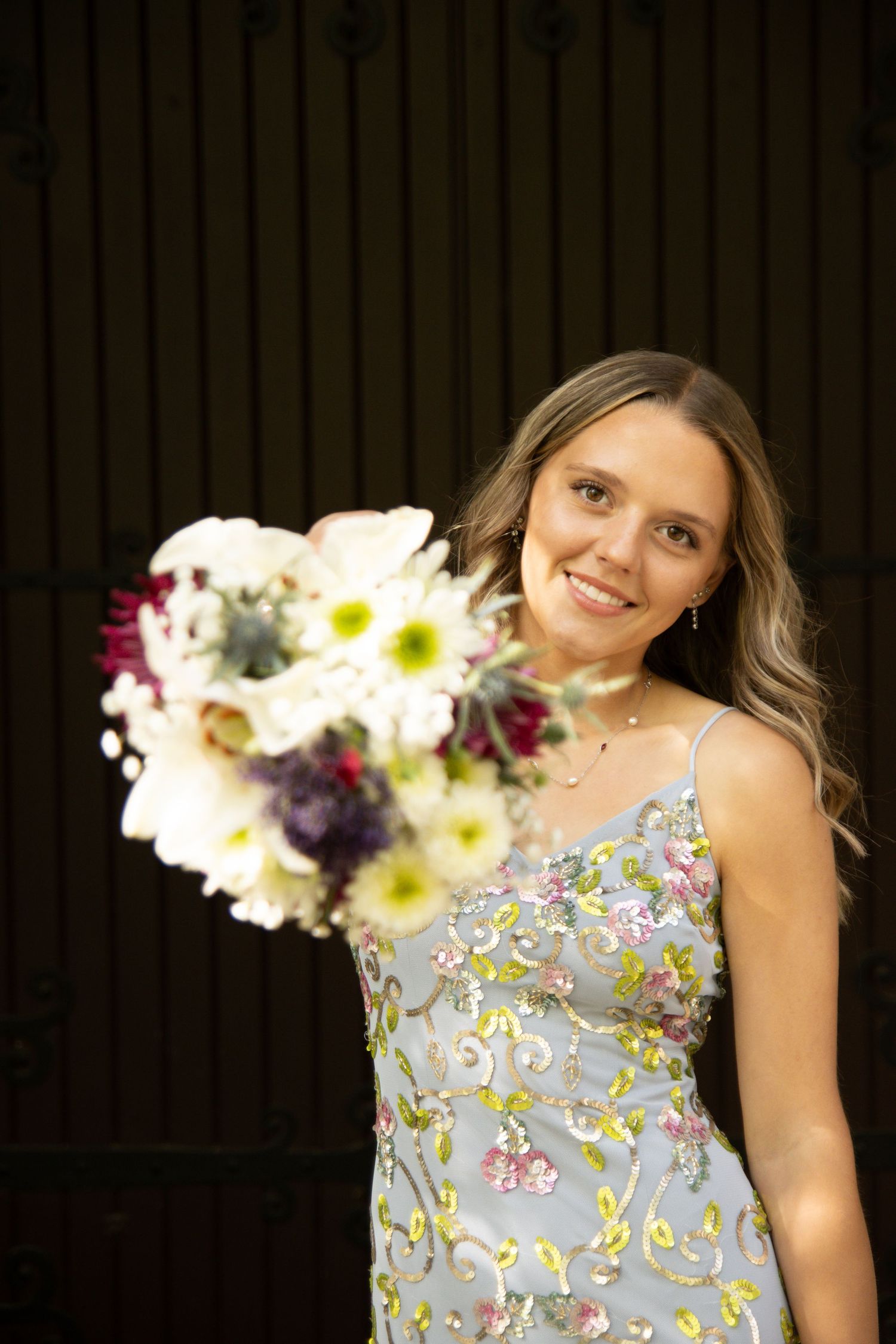 Smiling woman in floral embroidered blue dress holding a white and purple bouquet against dark wooden background.
