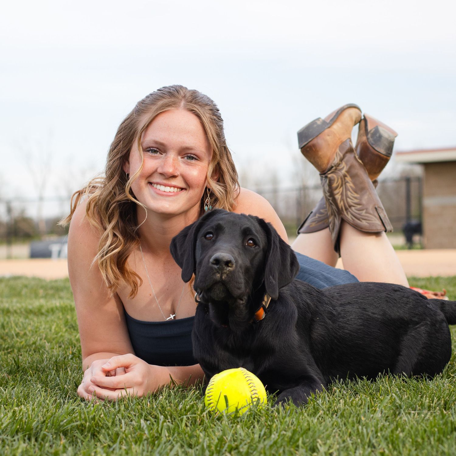 Young blonde woman smiling while lying on grass with a black Labrador dog and a yellow tennis ball outdoors.