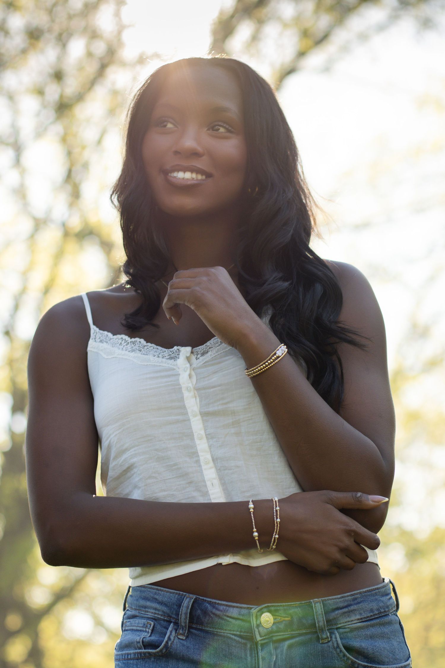 Smiling young Black woman in white lace cami and jeans, wearing gold bracelets, posing outdoors with sunlit trees behind her.