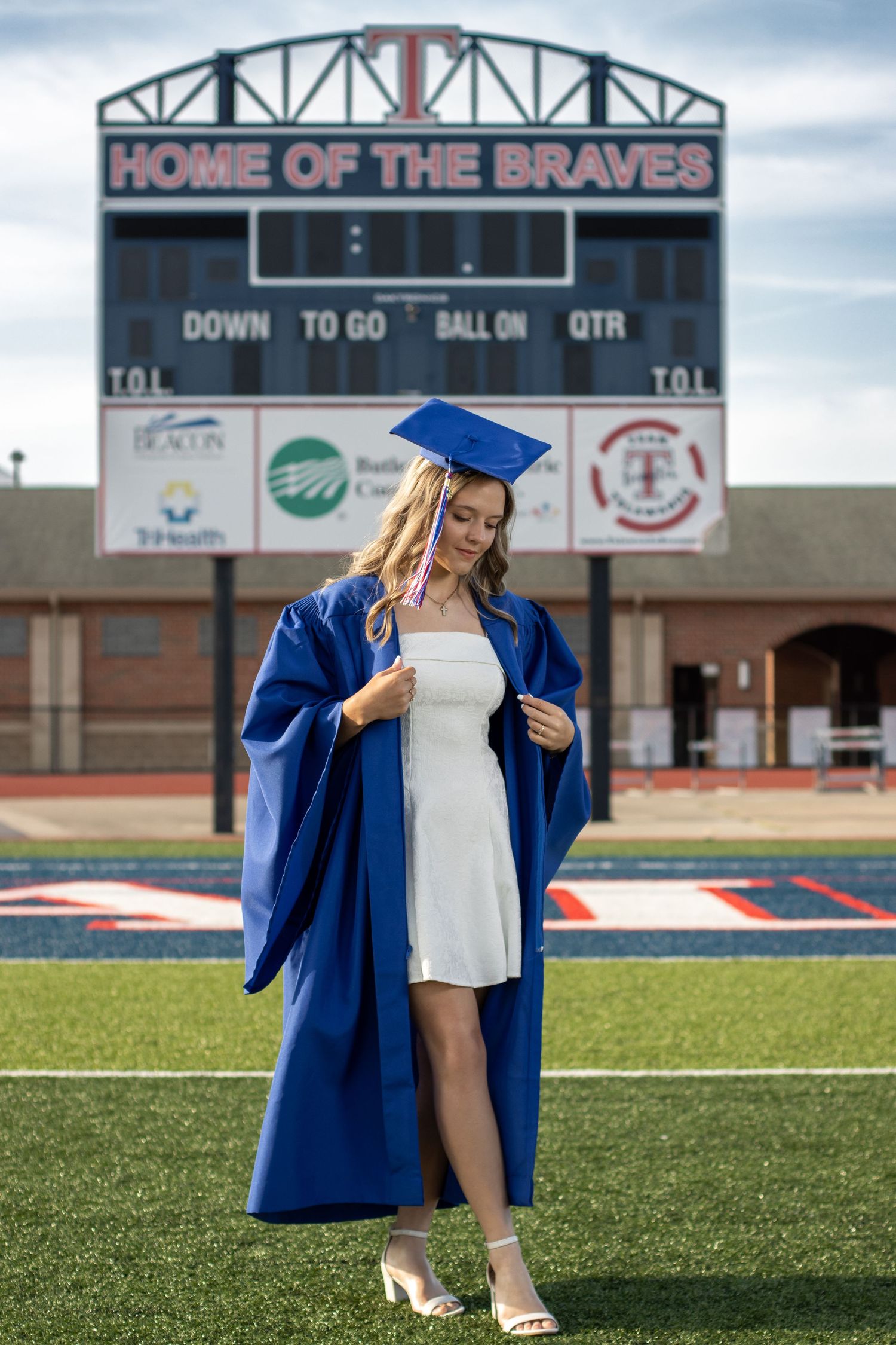 A young woman in a blue graduation cap and gown poses on a football field in front of a Home of the Braves scoreboard.