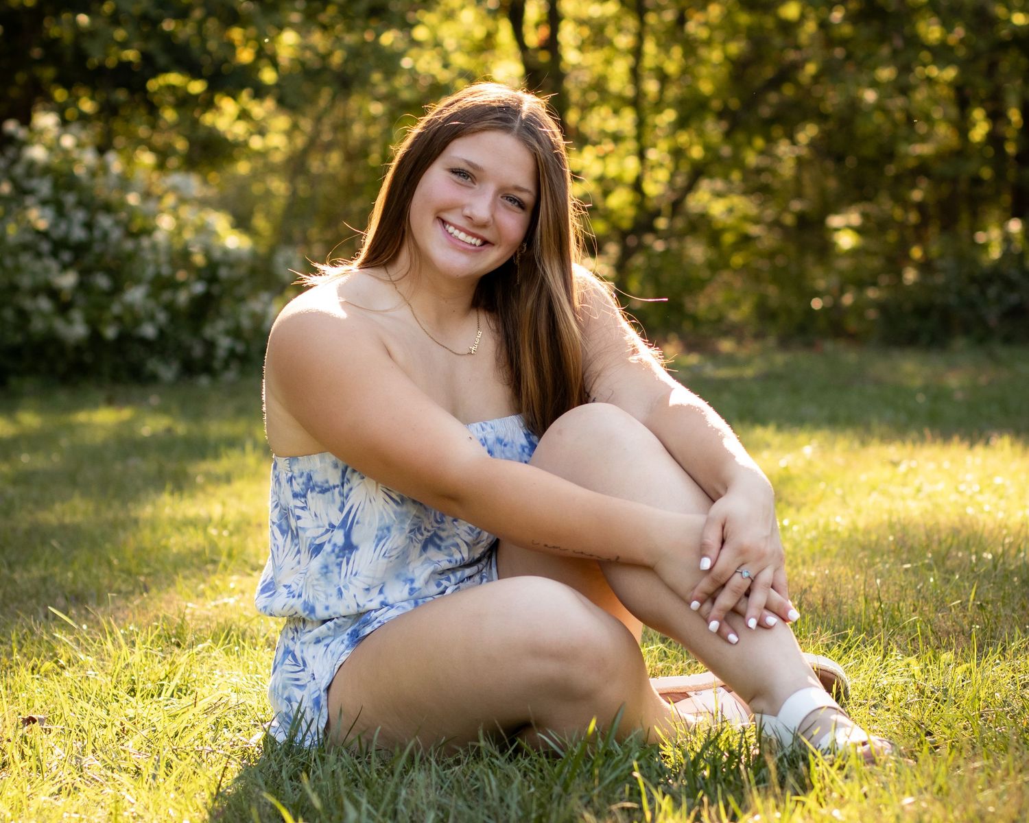 Person in floral dress sits casually on grass in sunny outdoor setting.