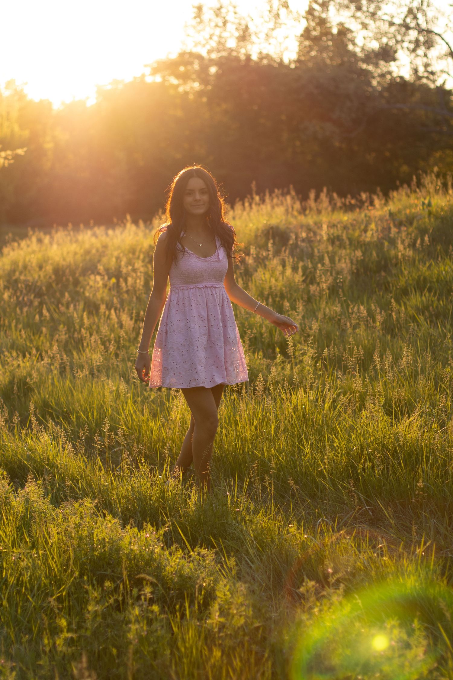 Young woman in white dress walking through golden sunlit meadow at sunset with warm lens flare.