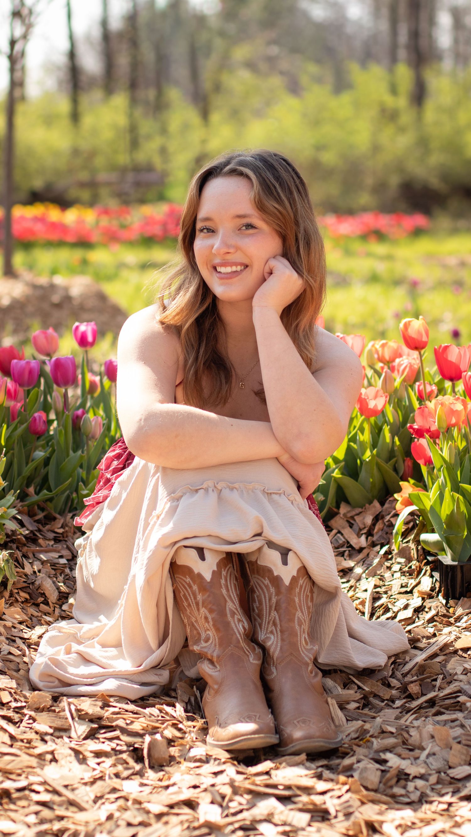 Smiling young woman in cream dress and cowboy boots sitting among colorful tulips in a sunny spring garden.