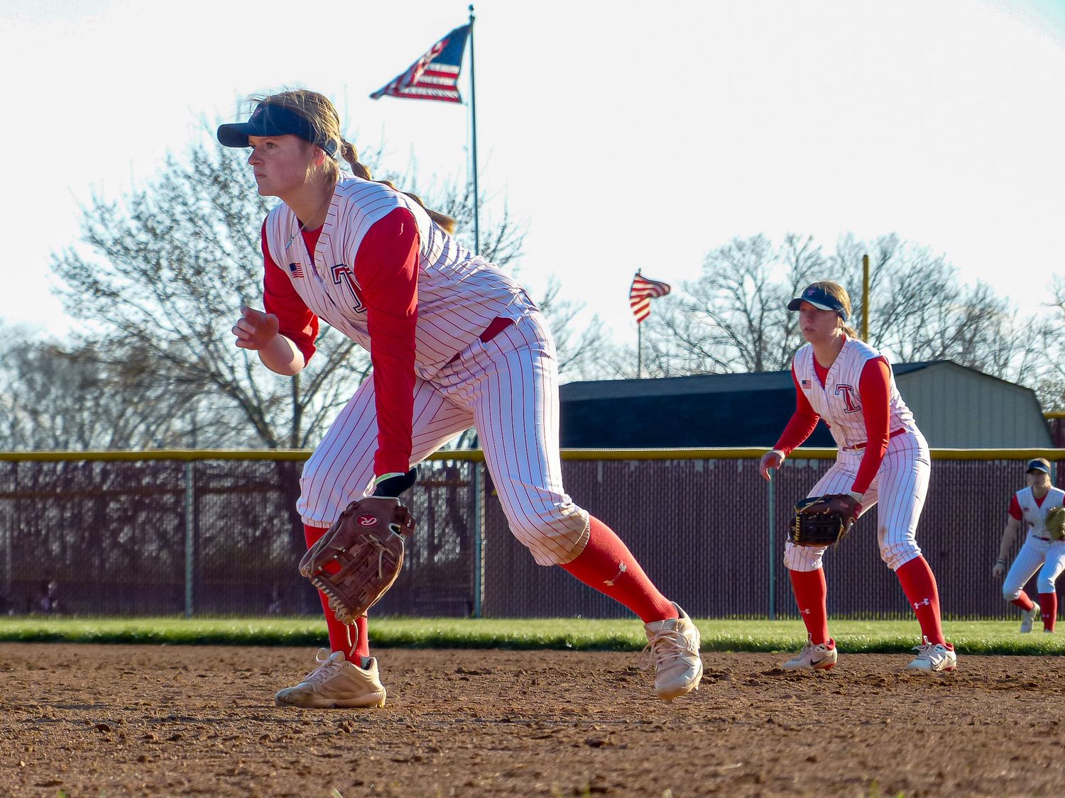 Baseball pitcher in red and white uniform follows through on delivery motion with American flag waving in background.