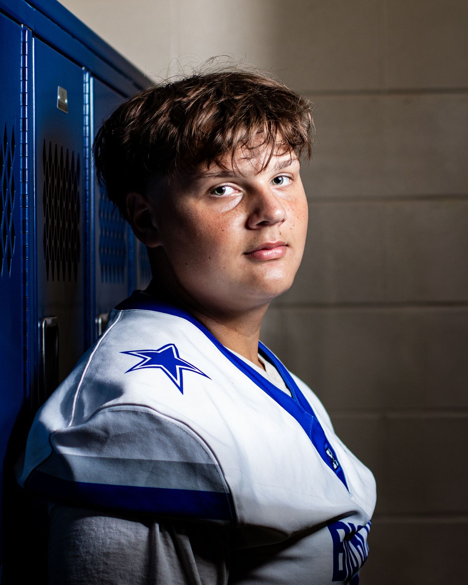 Young male athlete in white and blue football jersey leaning against blue lockers for a senior portrait photo.