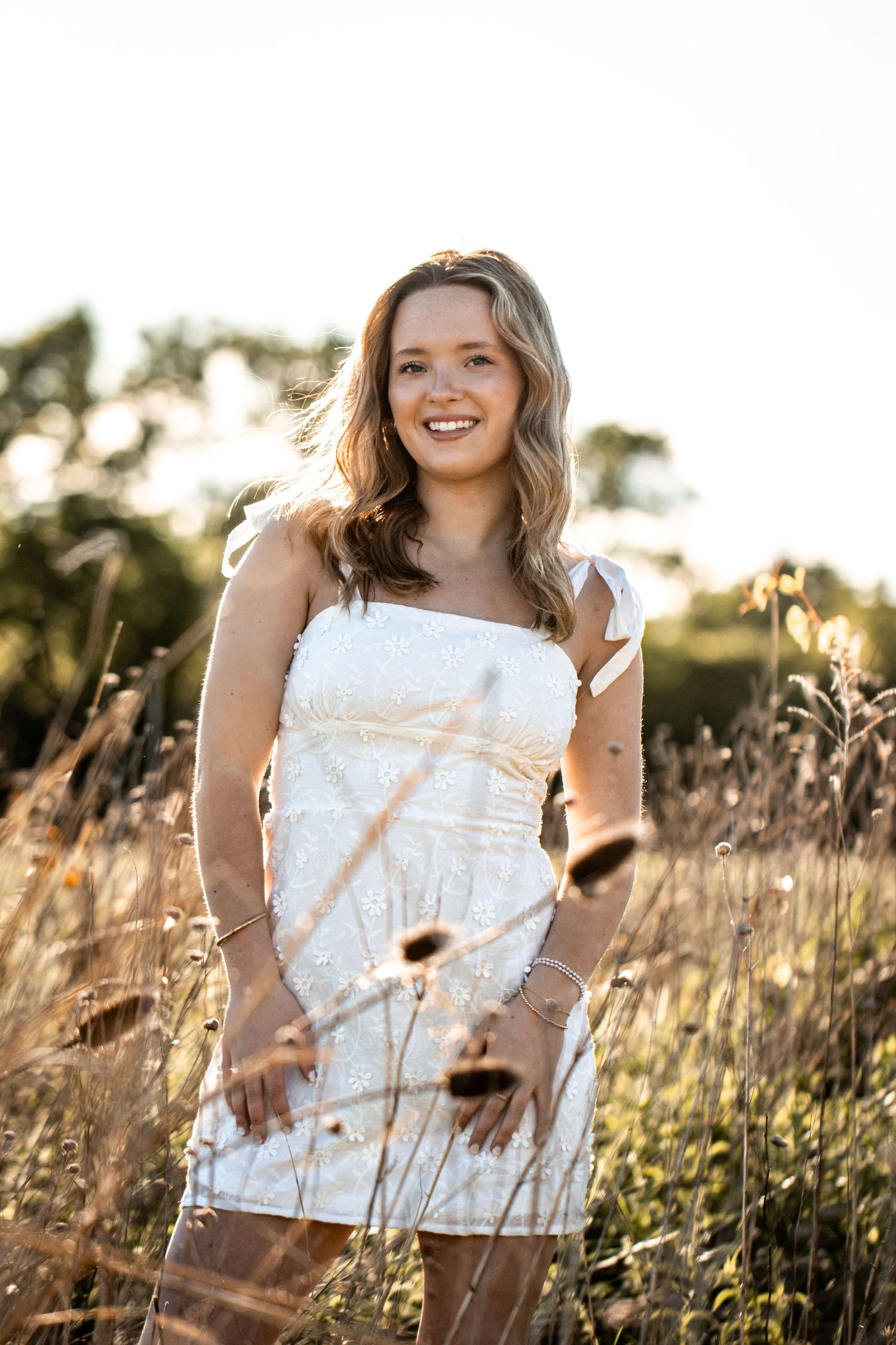 Young woman in white mini dress smiling in golden sunlit field with tall grasses and wildflowers.