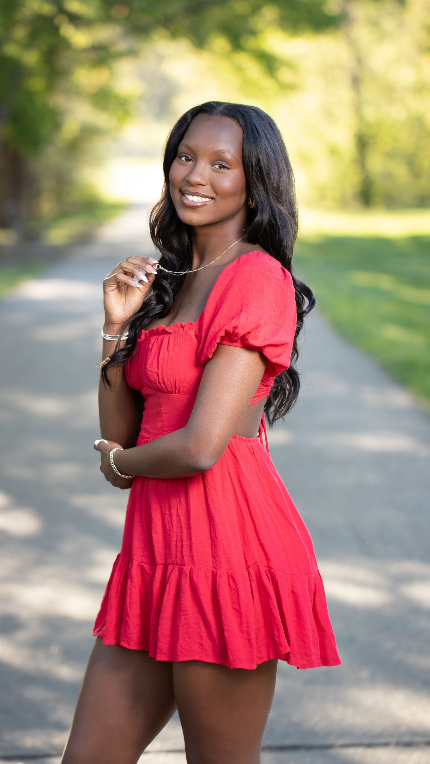 Young woman in a red mini dress smiling outdoors on a tree-lined path, posing for a portrait photo.