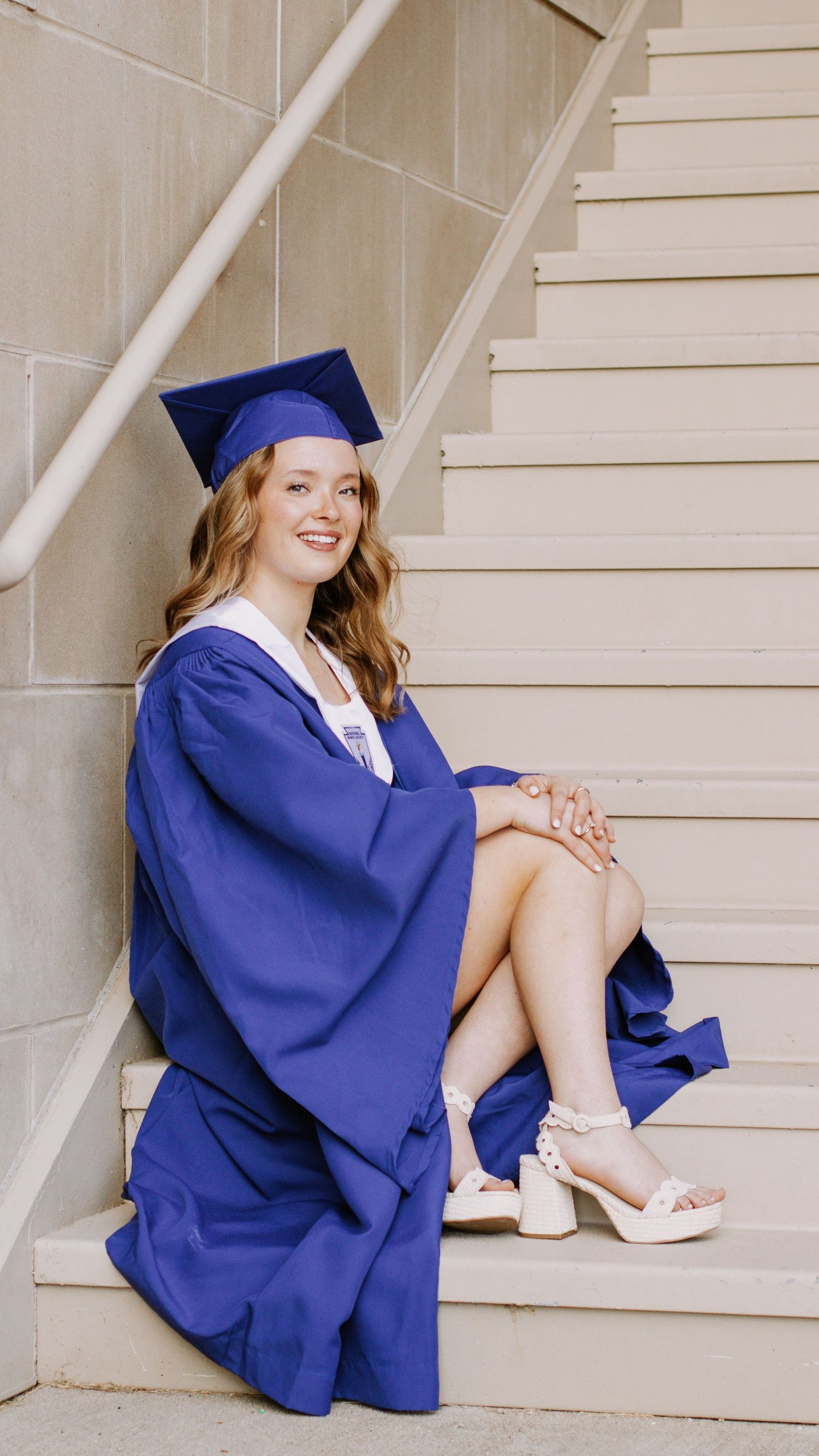 Young woman in blue graduation cap and gown sitting on stairs, smiling, wearing white platform heels.
