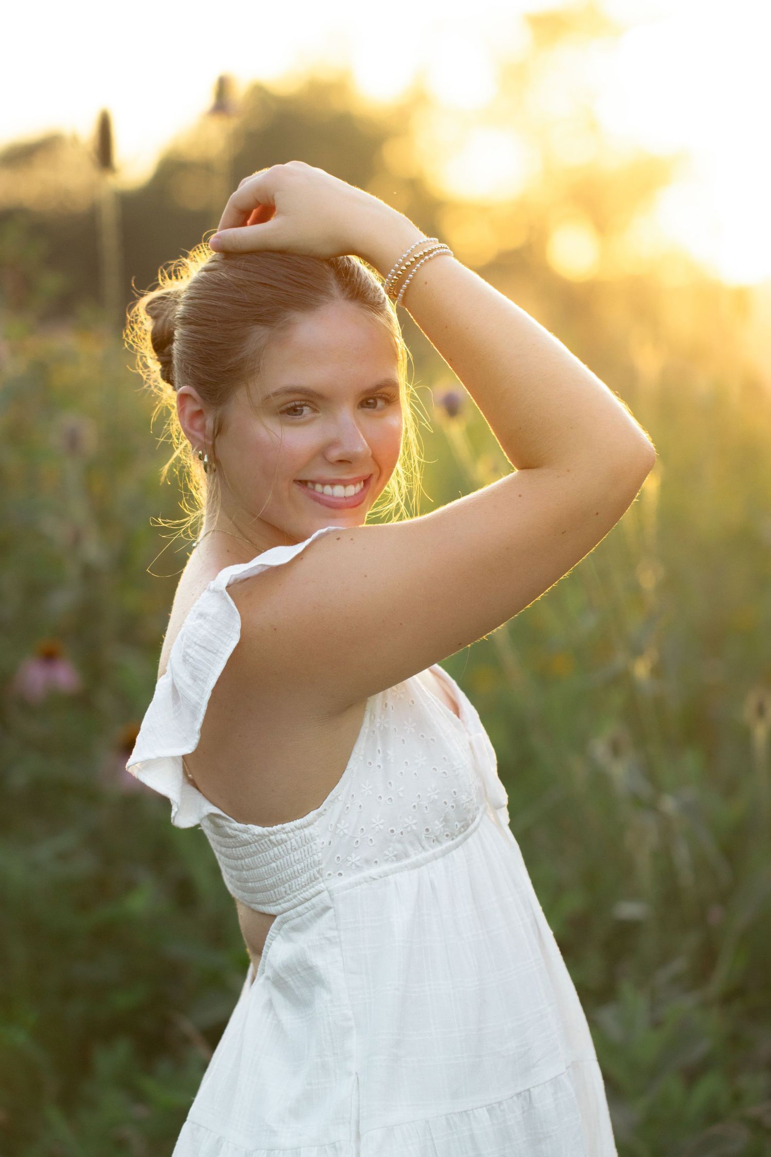 Young woman in white dress smiling in golden sunset field, hand raised to hair, wearing gold bracelet.