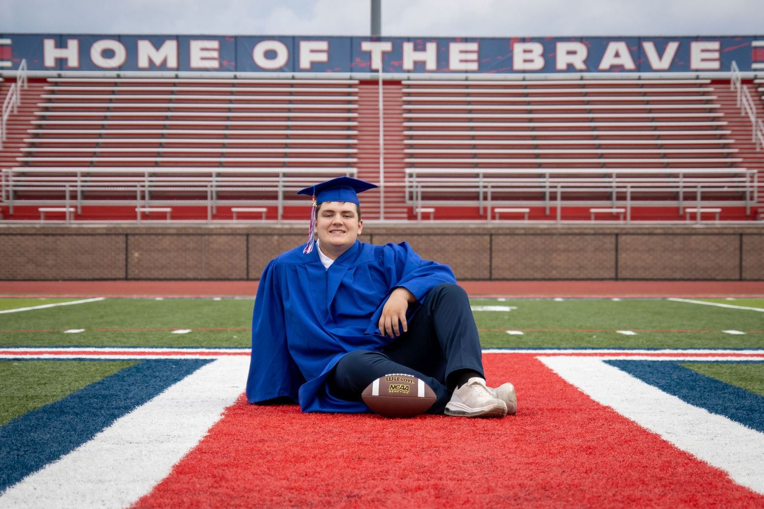 Graduate in blue cap and gown sits on football field turf holding a football, with Home of the Brave sign behind.