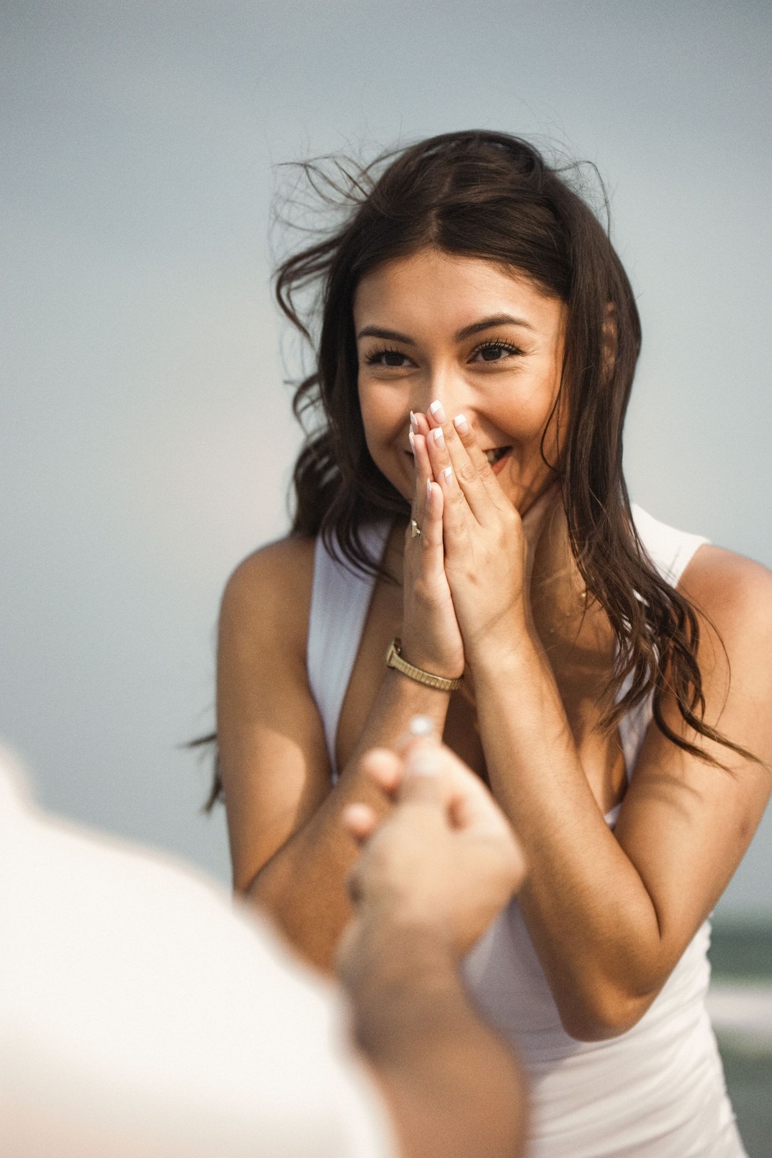 Surprised woman in white top covers mouth with hands, smiling joyfully during a romantic proposal outdoors.