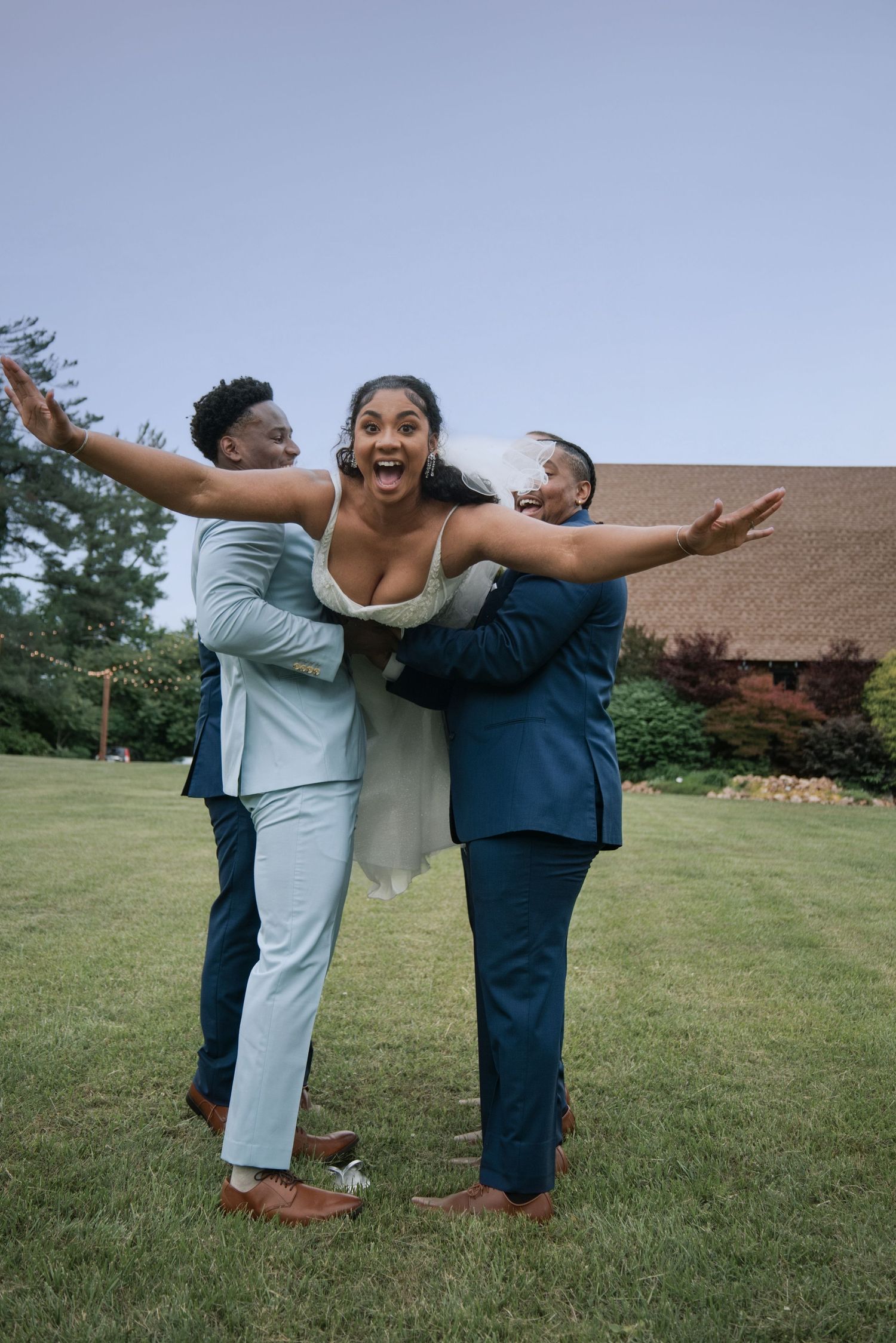 Bride in white dress laughs joyfully as two groomsmen in blue suits playfully lift her outdoors.