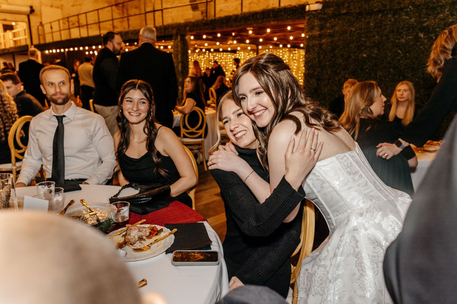 Two women hugging and smiling at a wedding reception dinner, with guests and string lights in the background.