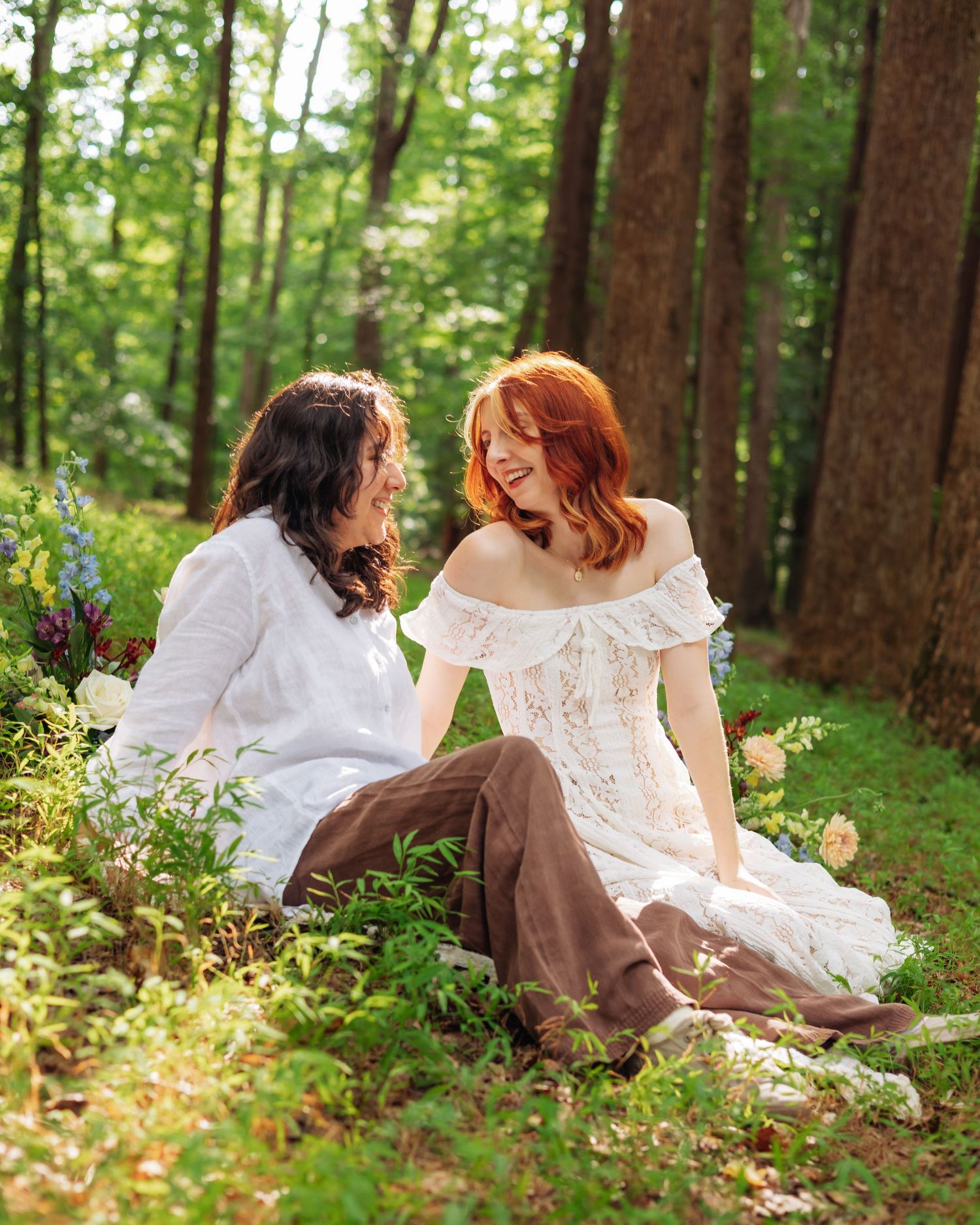 A couple in white lace and brown pants sit laughing in a lush green forest surrounded by wildflowers.
