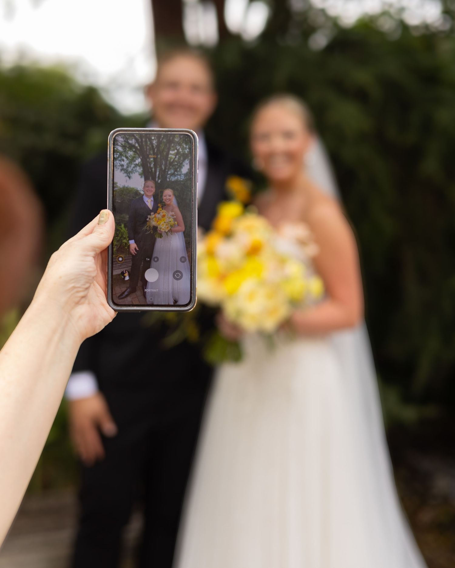 A hand holds a smartphone capturing a bride and groom posing outdoors, with the couple blurred in the background.