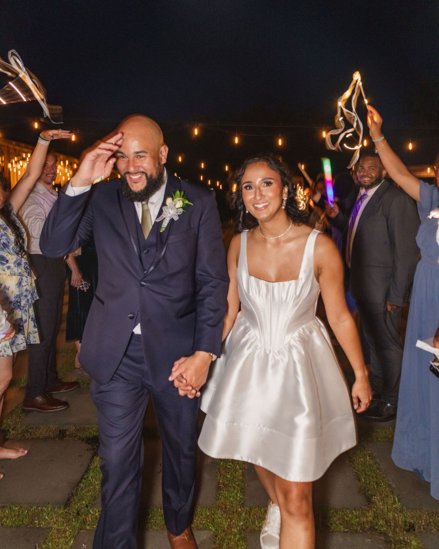 A joyful newlywed couple walks hand-in-hand at their wedding reception, surrounded by guests holding sparklers at night.