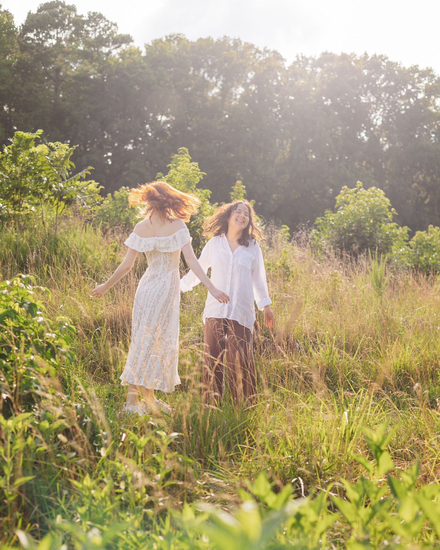 Two women in white dresses hold hands running through a sunlit meadow with lush green trees in the background.