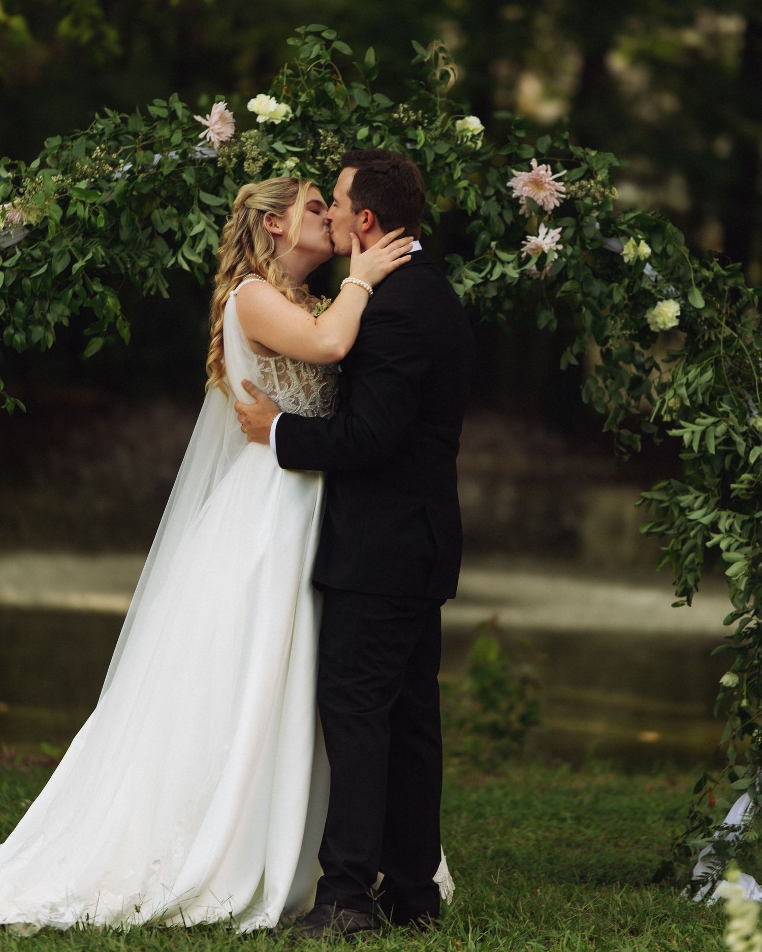 Bride and groom share their first kiss at an outdoor wedding ceremony with a lush green floral arch backdrop.