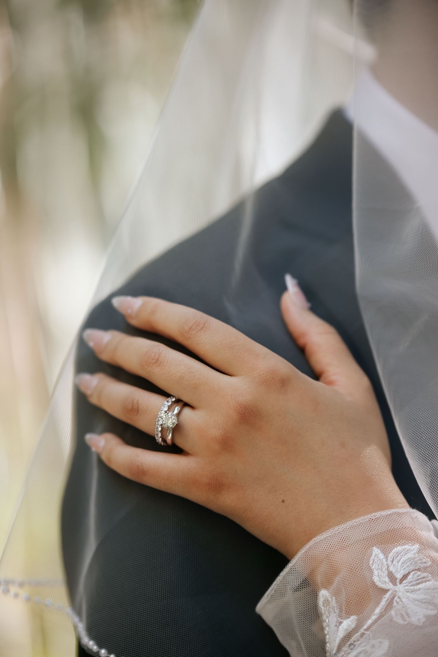 Close-up of a bride's hand with diamond engagement ring and wedding band resting on groom's dark suit jacket.