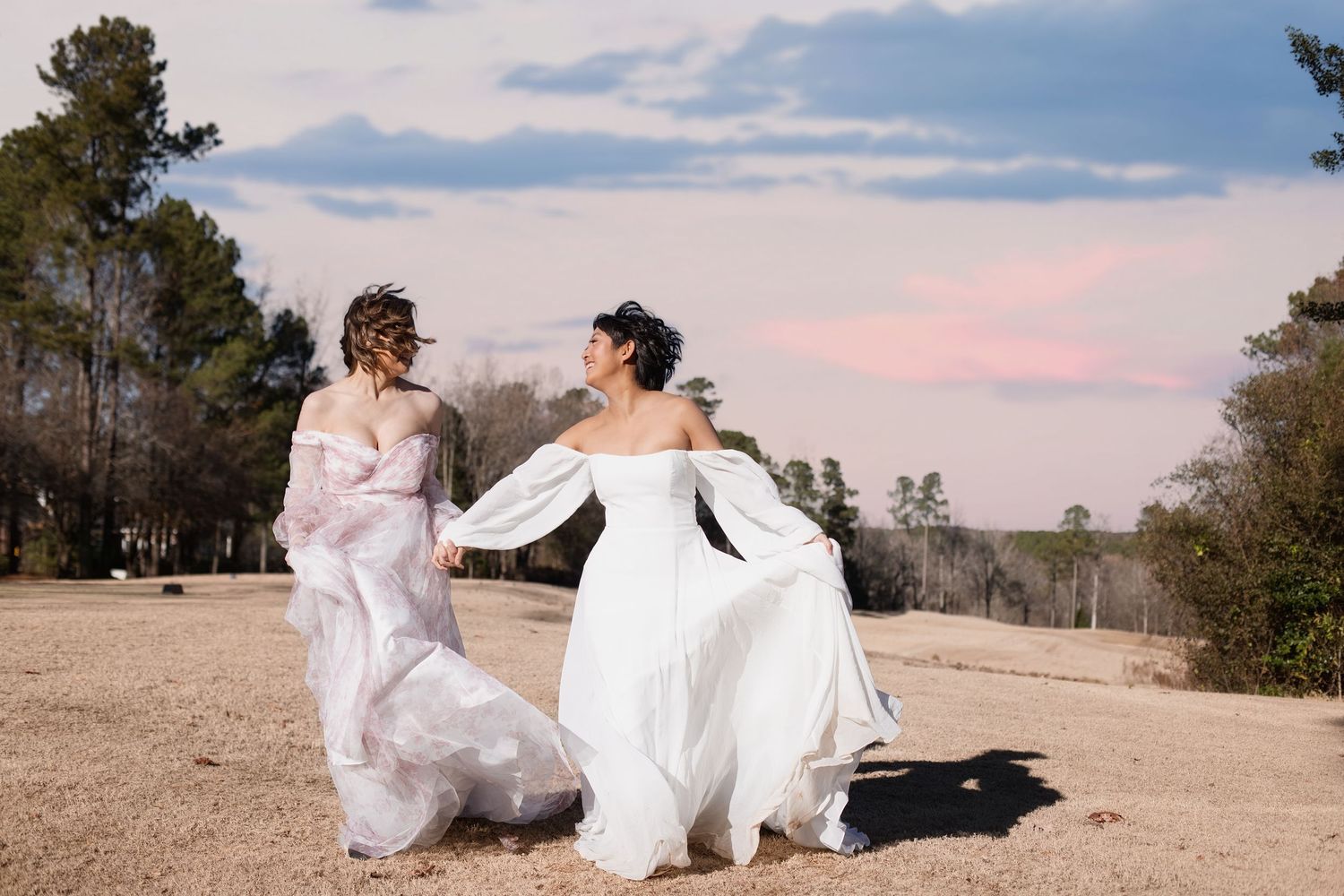 Two brides in white wedding gowns hold hands walking outdoors at sunset with trees and pastel sky behind them.