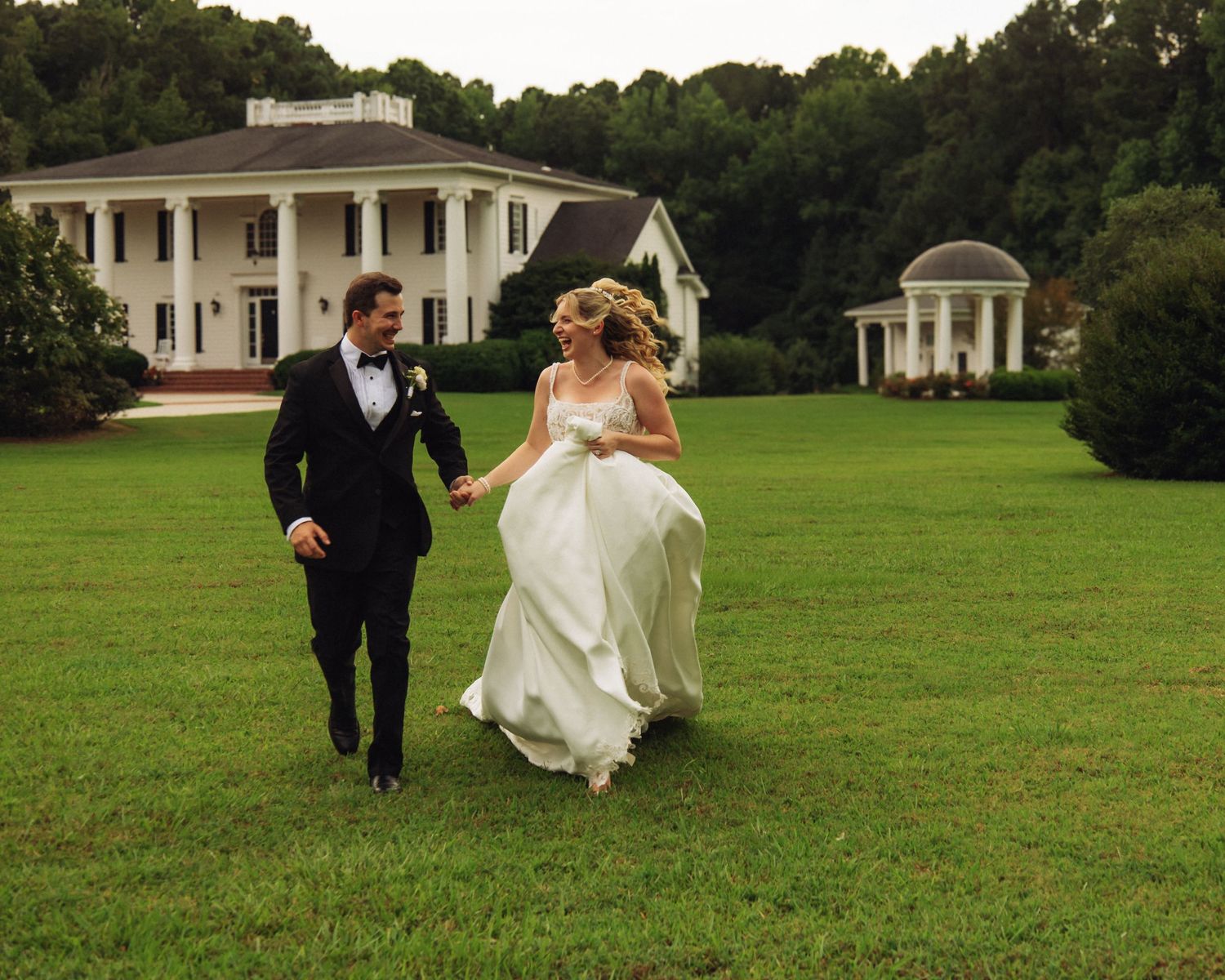 Bride and groom holding hands running across green lawn in front of elegant white Southern mansion on wedding day.