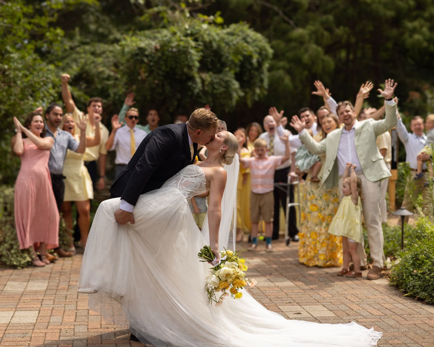 Bride and groom share a romantic dip kiss at outdoor wedding ceremony while guests cheer and wave behind them.