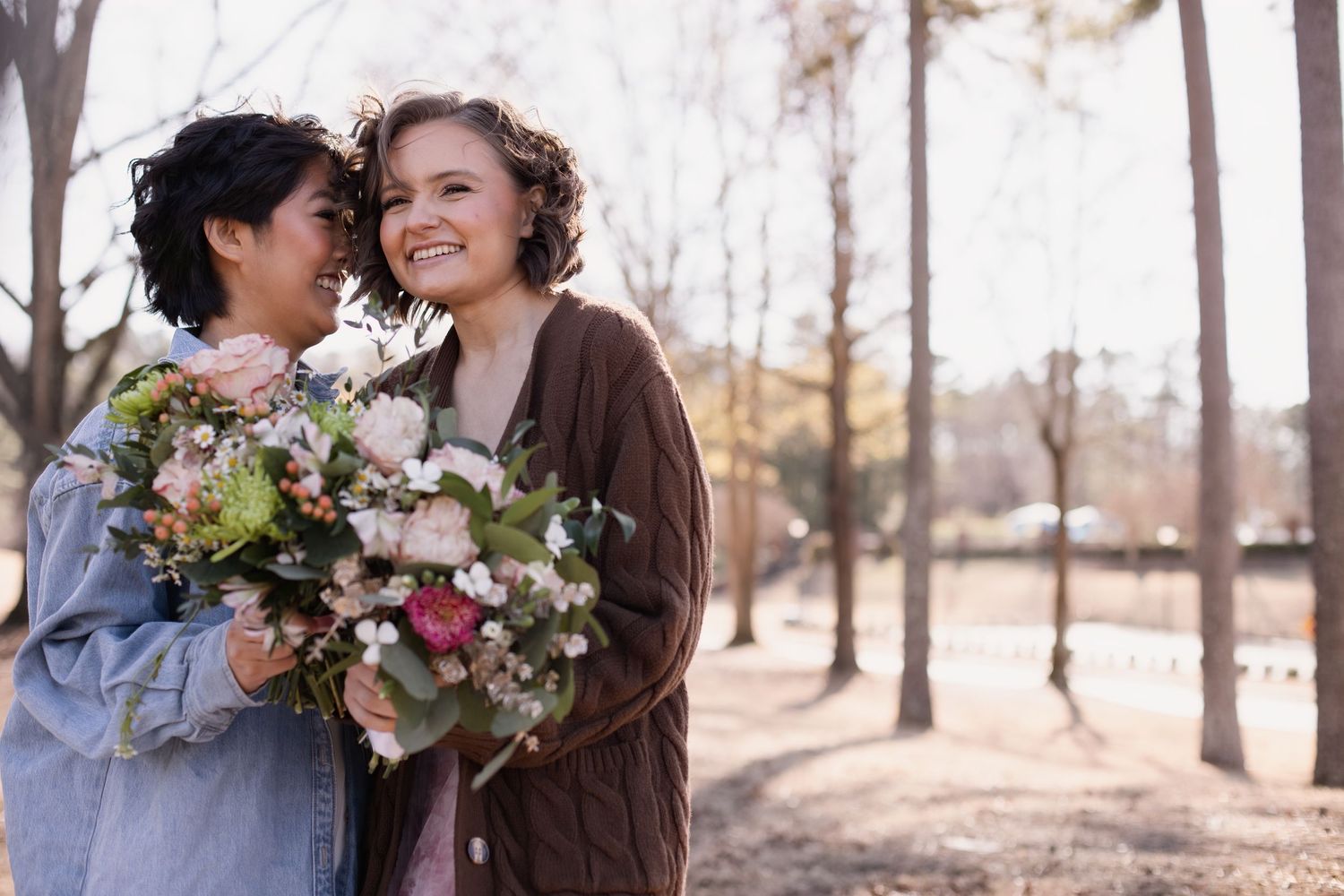Two women smiling and holding bouquets of flowers outdoors in a park during autumn.