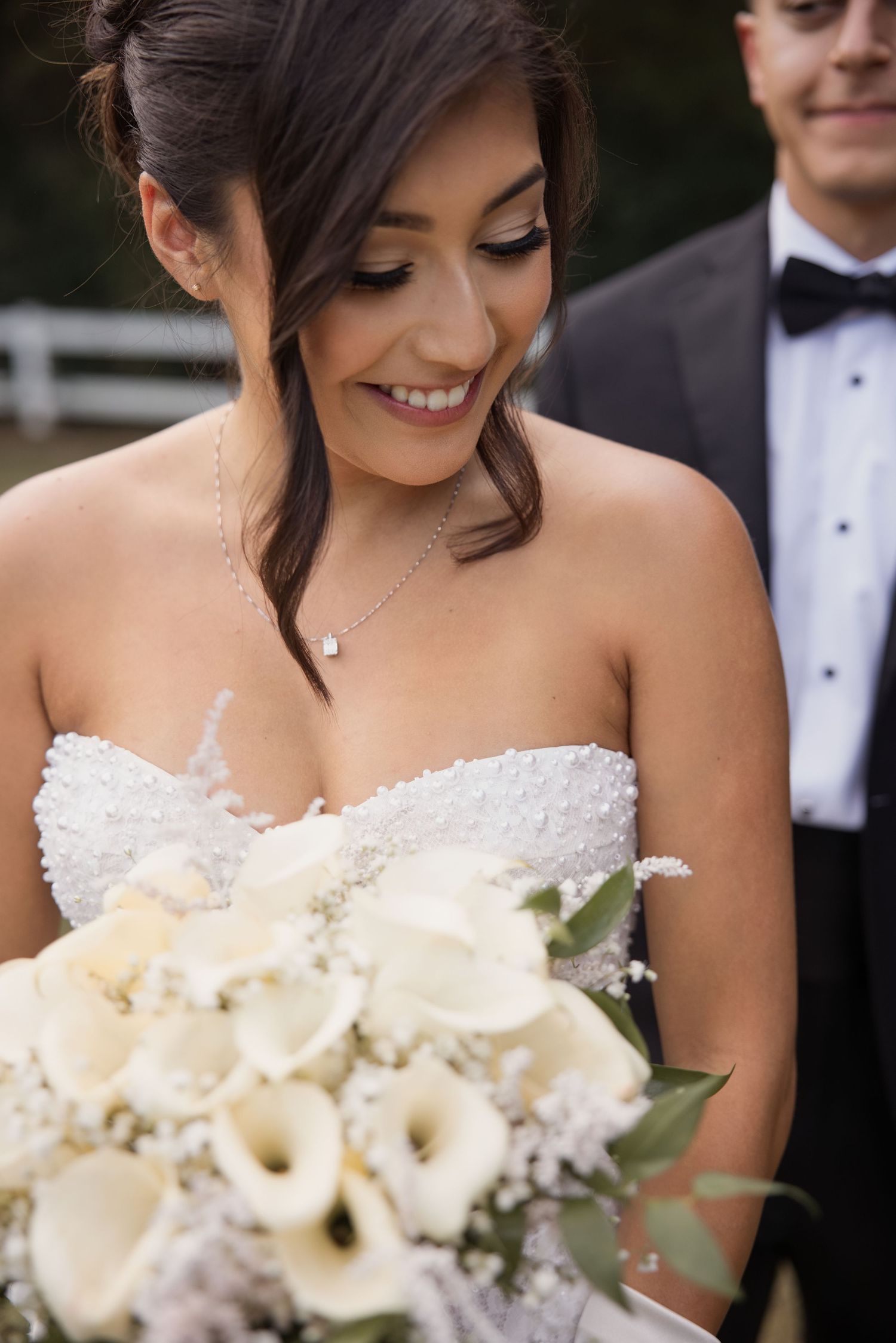Smiling bride in strapless beaded gown holds white calla lily bouquet, with groom in tuxedo visible behind her.