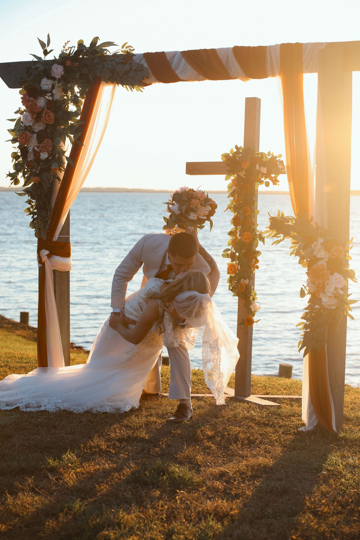 Couple sharing a romantic dip kiss under a floral arch at a waterfront wedding ceremony during golden hour sunset.