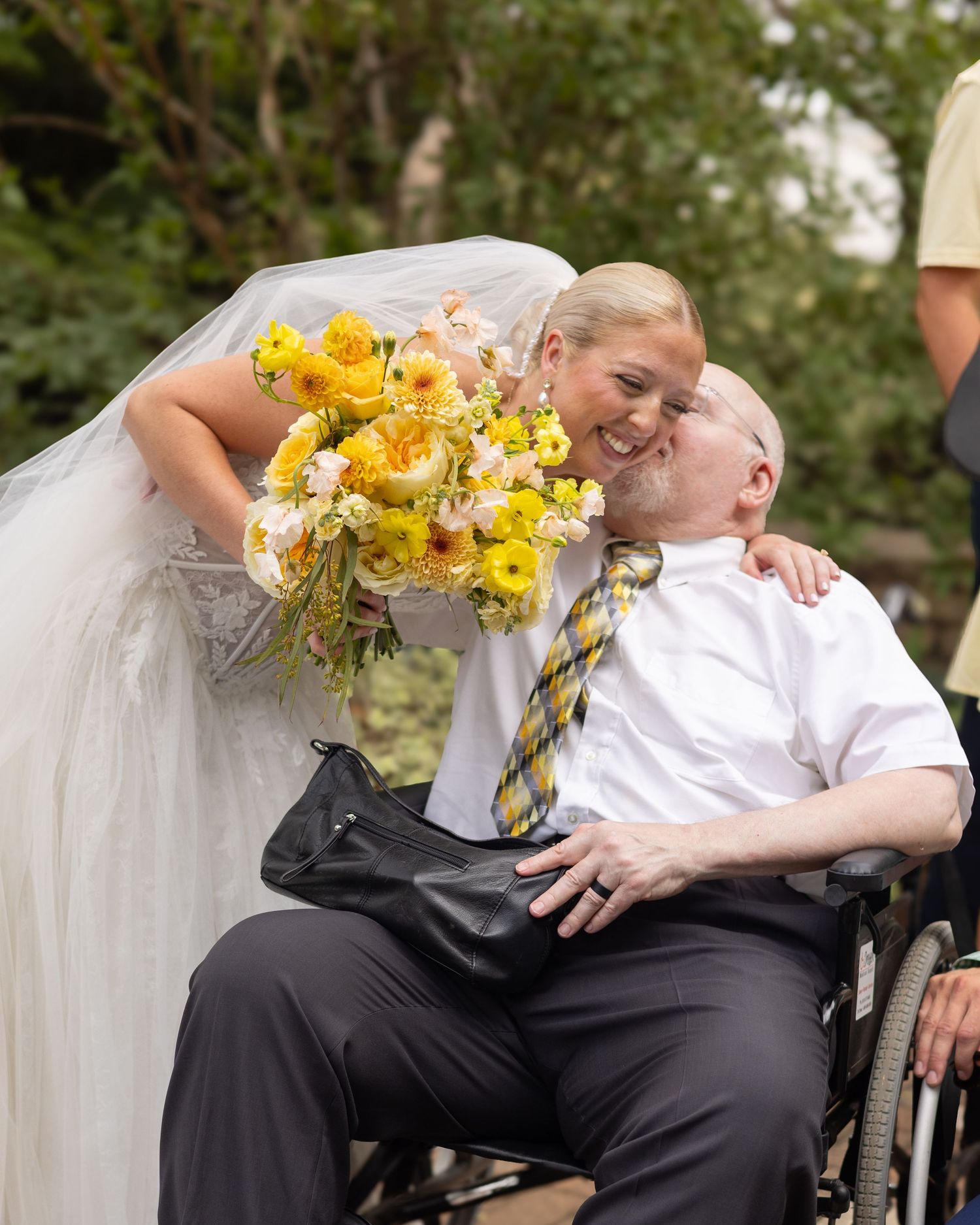Bride in white gown holds yellow bouquet while leaning down to kiss man in wheelchair wearing striped tie outdoors.
