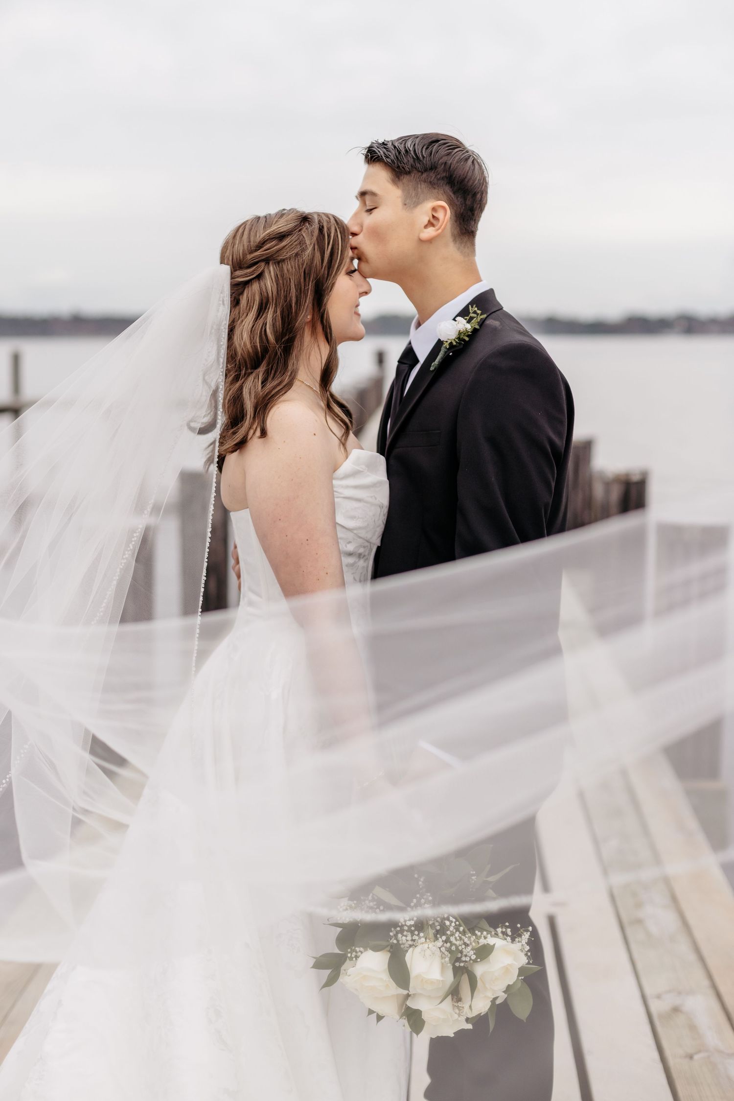 Bride and groom share a romantic moment on a waterfront dock, with the wedding veil flowing gracefully in the breeze.