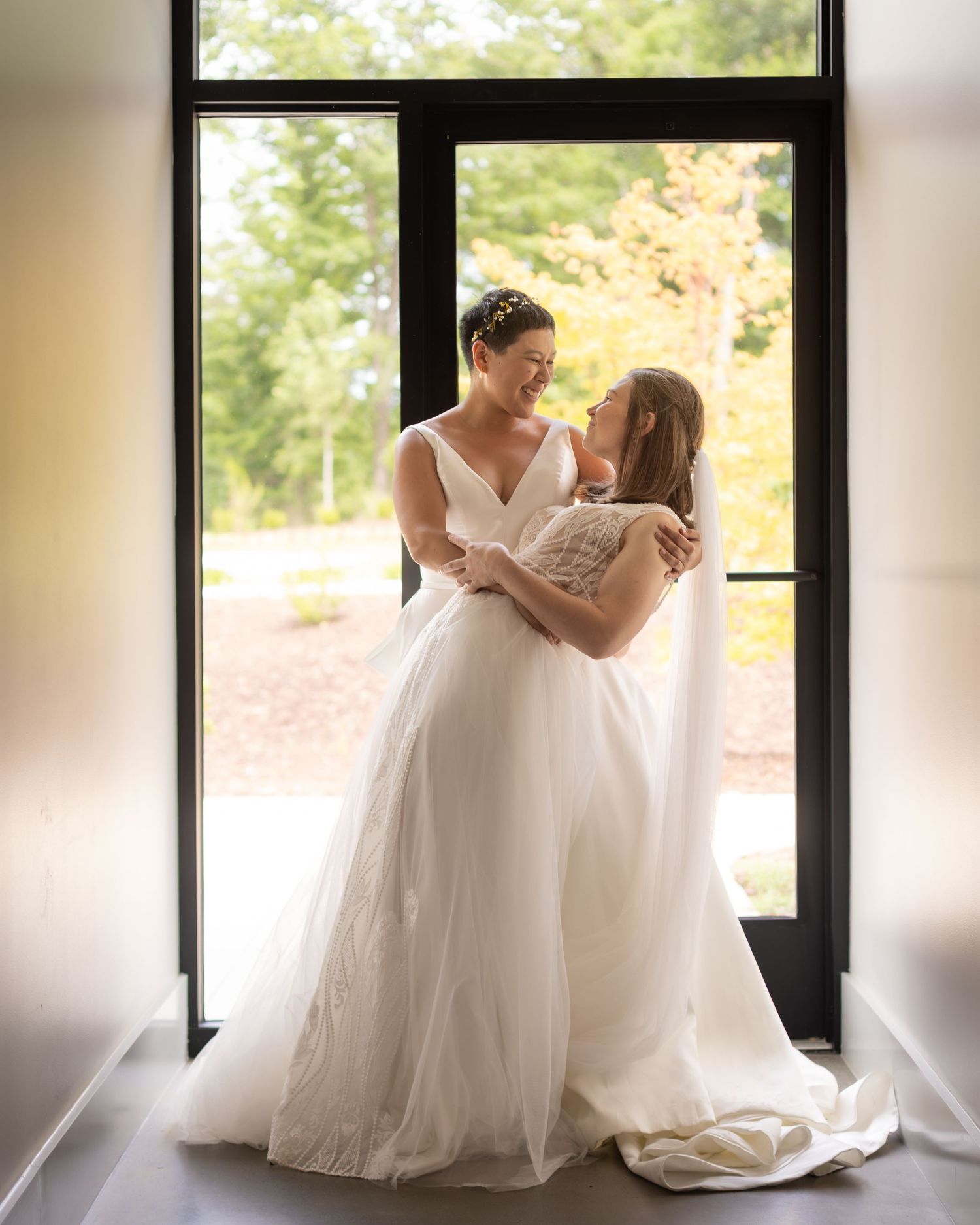 Two brides in white wedding gowns embrace and smile at each other in a sunlit doorway surrounded by autumn trees.
