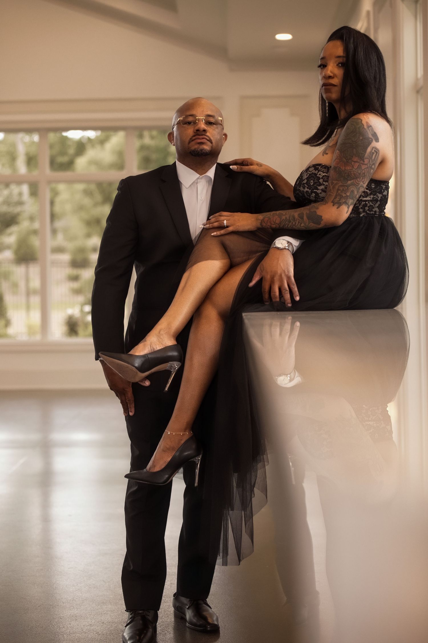A man in a black suit poses with a tattooed woman in an elegant black gown, leaning against a reflective surface.