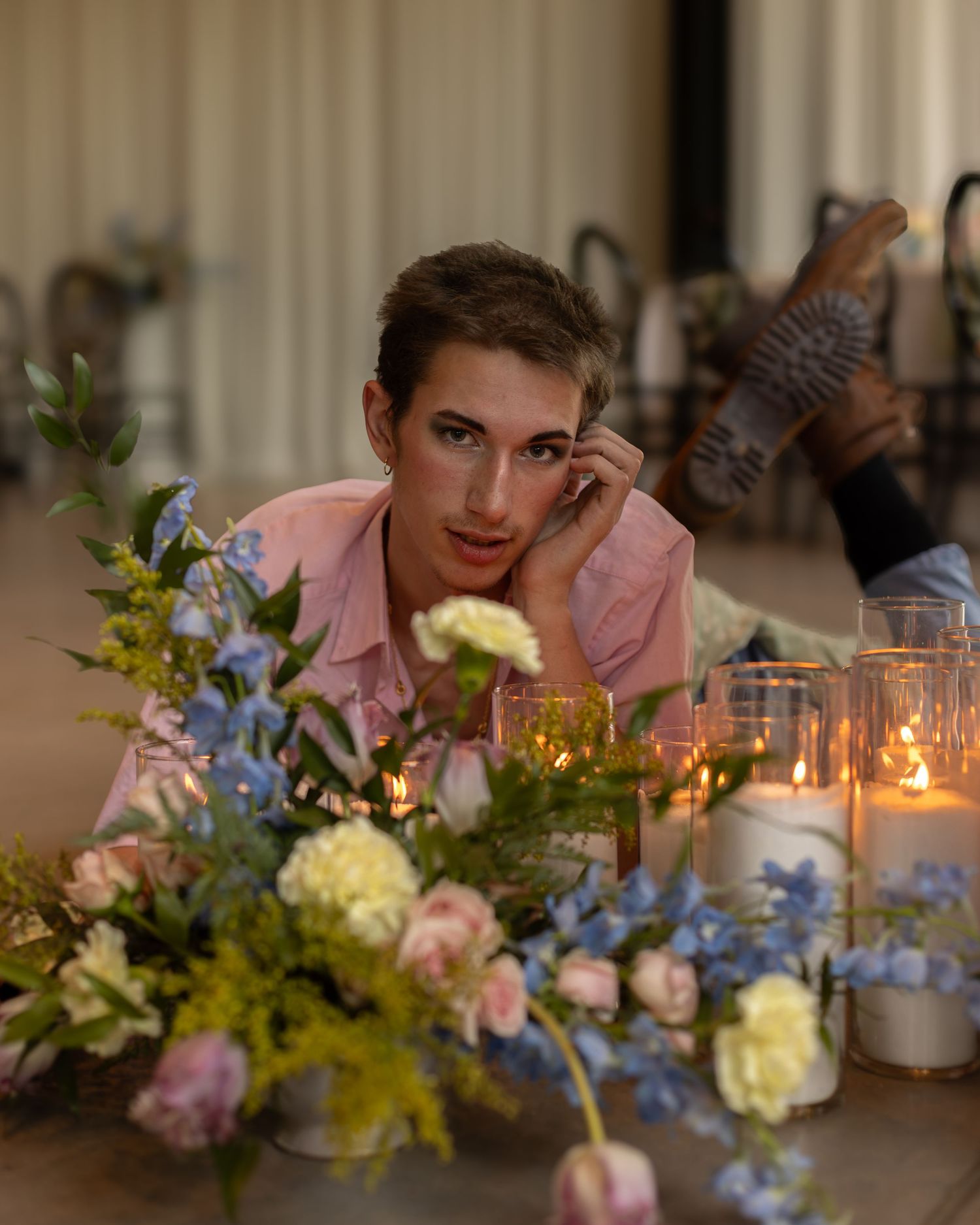 Person lying on floor surrounded by pink and blue flowers and glowing candles in glass holders.
