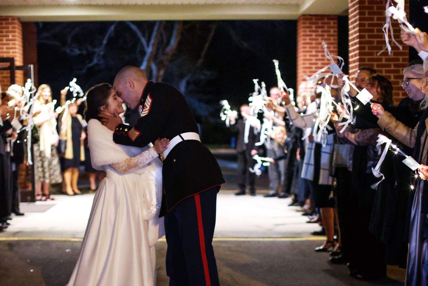 Military wedding couple kissing during sparkler send-off exit at night, surrounded by cheering guests.