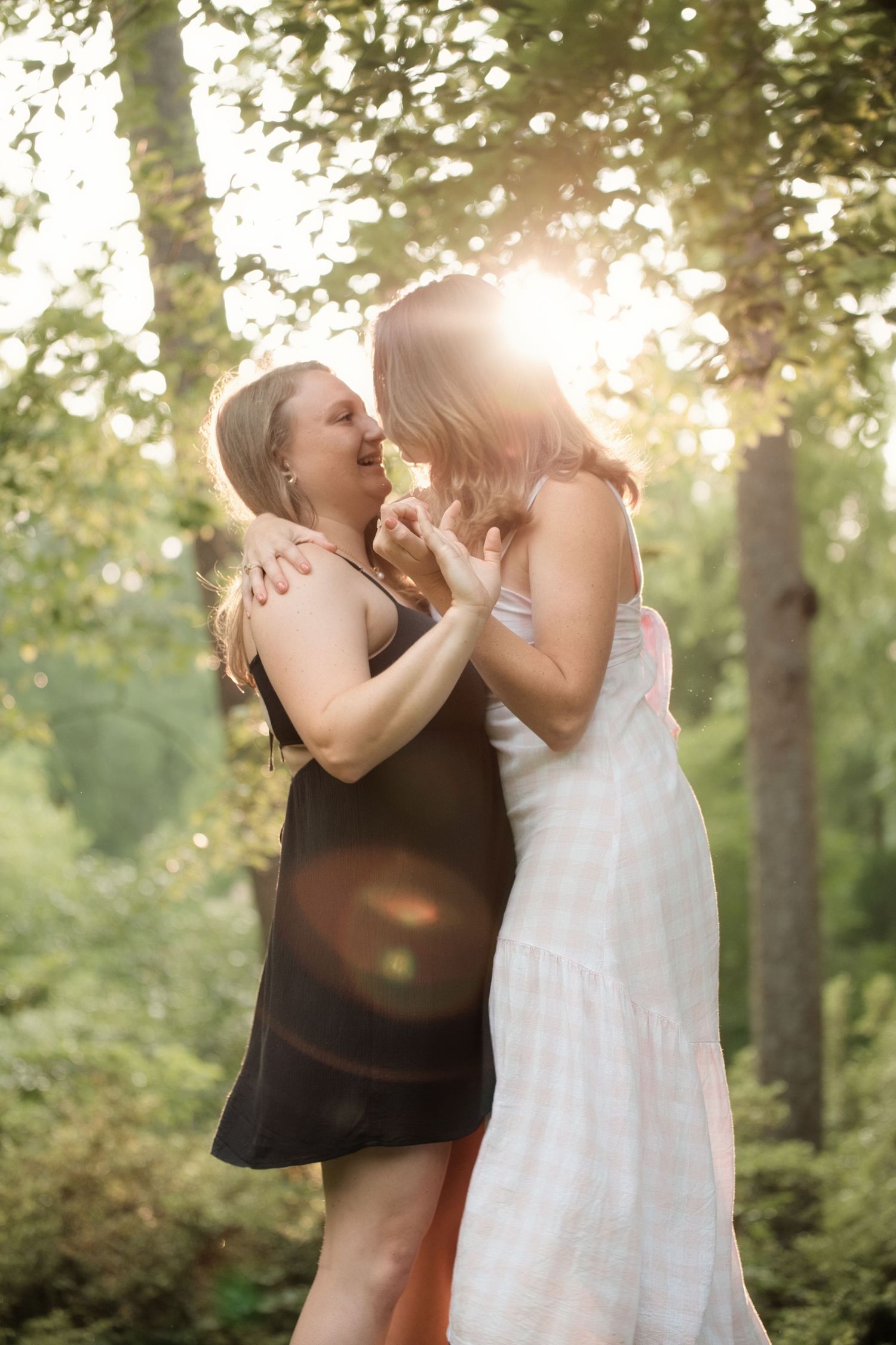 Two women in summer dresses laughing and embracing in a sunlit forest with golden backlight.