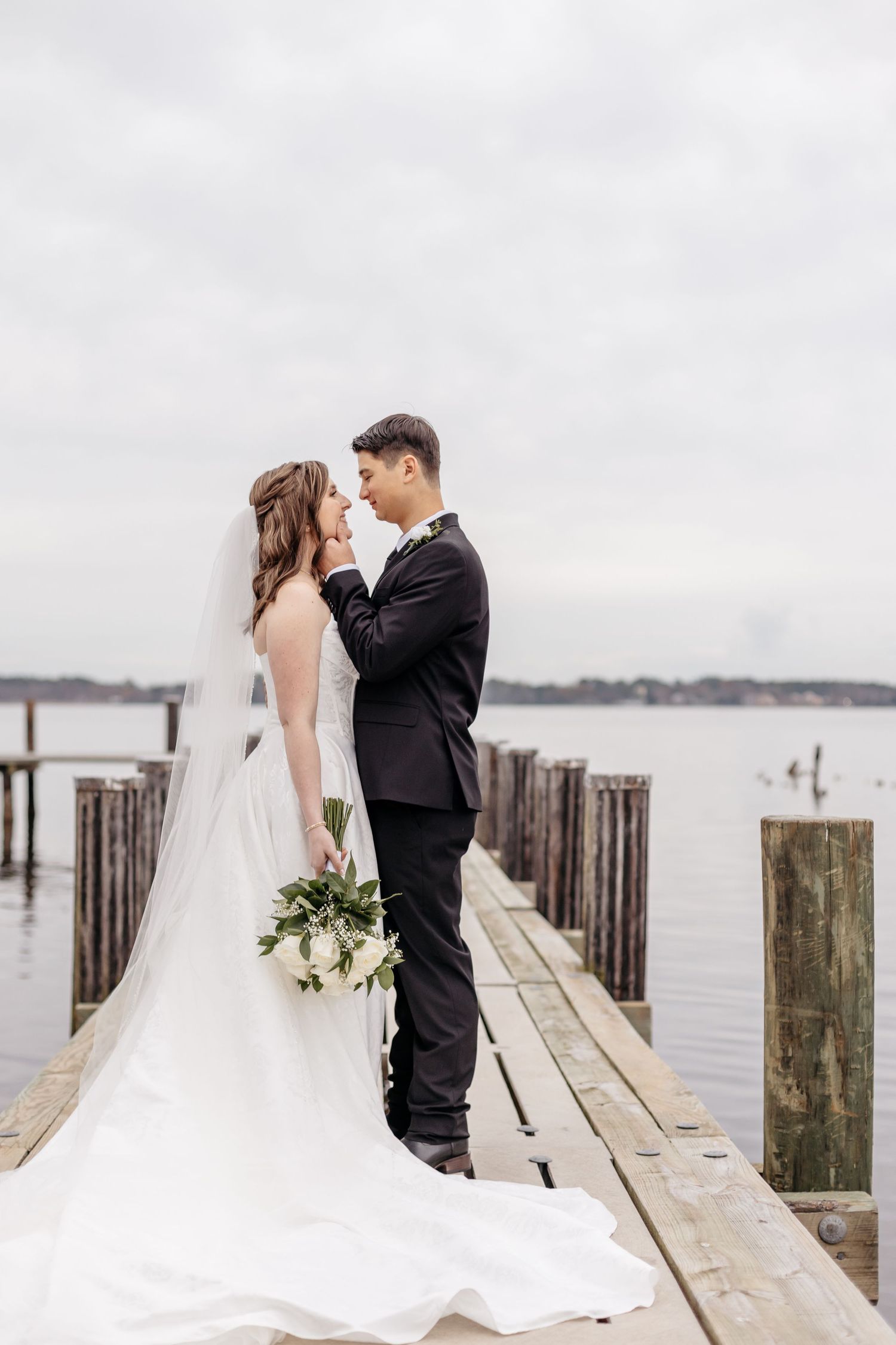 Bride and groom share a romantic moment on a wooden dock by the water, she in a flowing gown, he in a black suit.