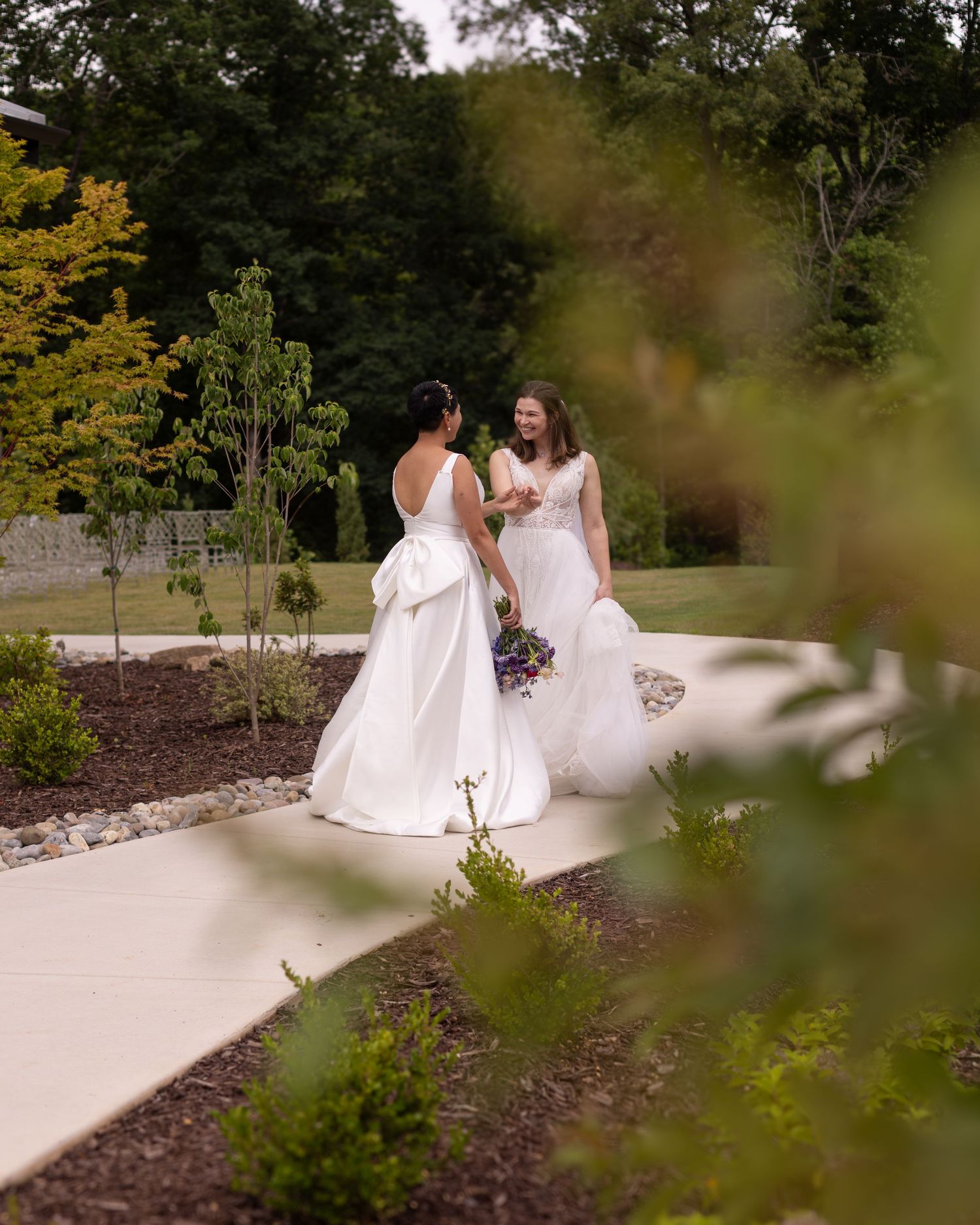 Two brides in white gowns share a joyful moment on a garden pathway surrounded by lush greenery.