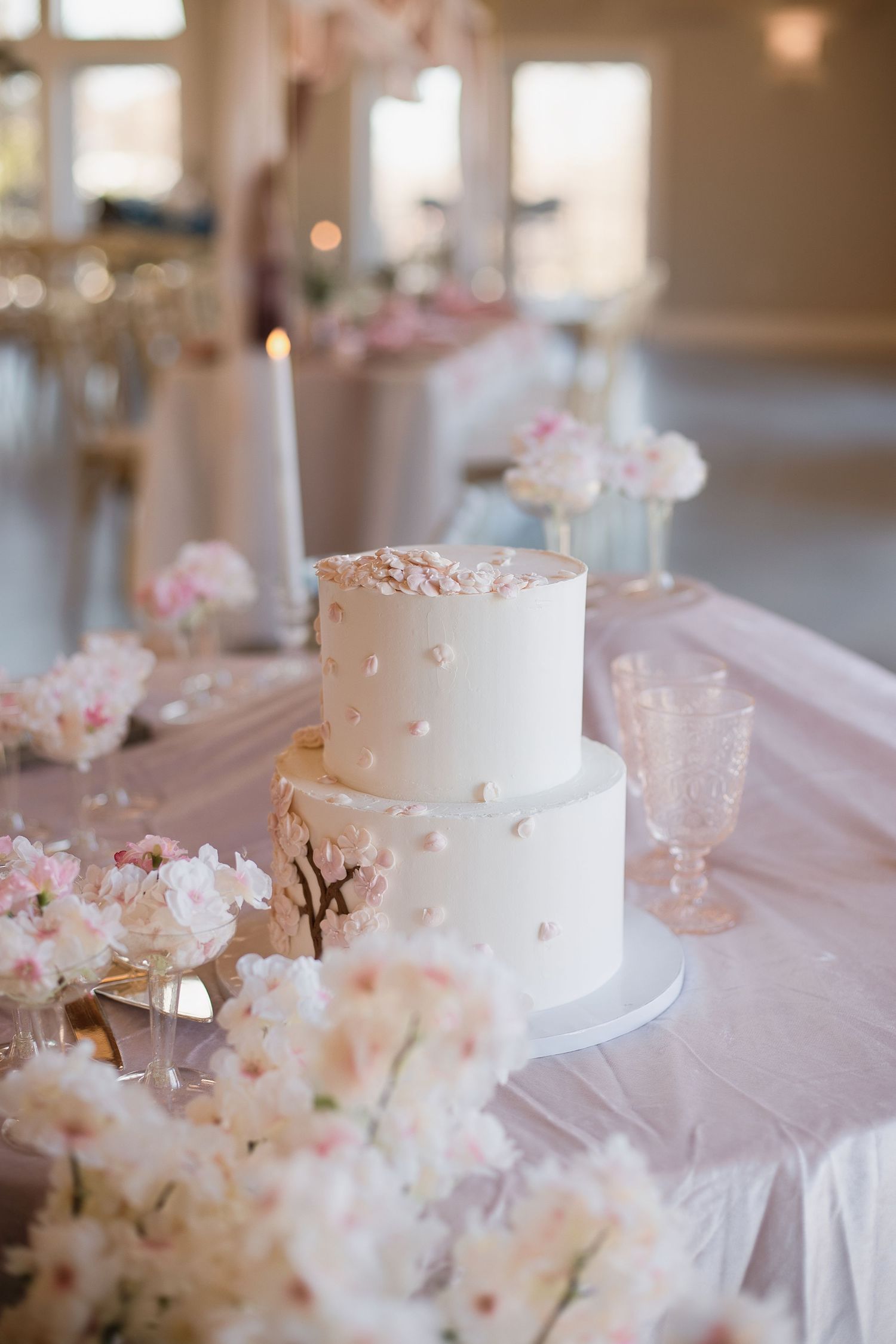 Elegant two-tier white wedding cake with pink floral details surrounded by cherry blossoms on a pink reception table.