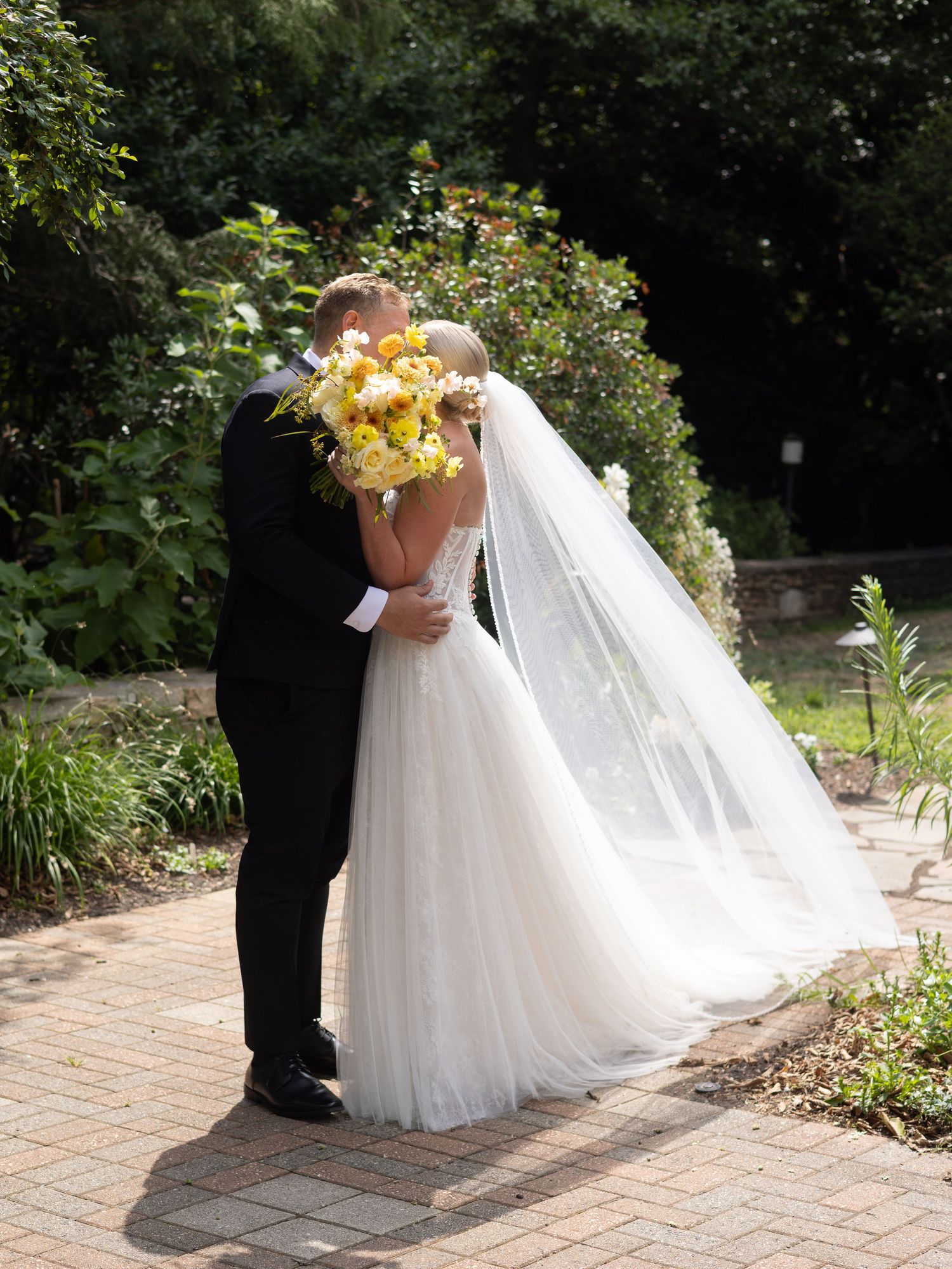 A bride and groom share a kiss on a brick path, surrounded by lush greenery, with a flowing veil and yellow bouquet.