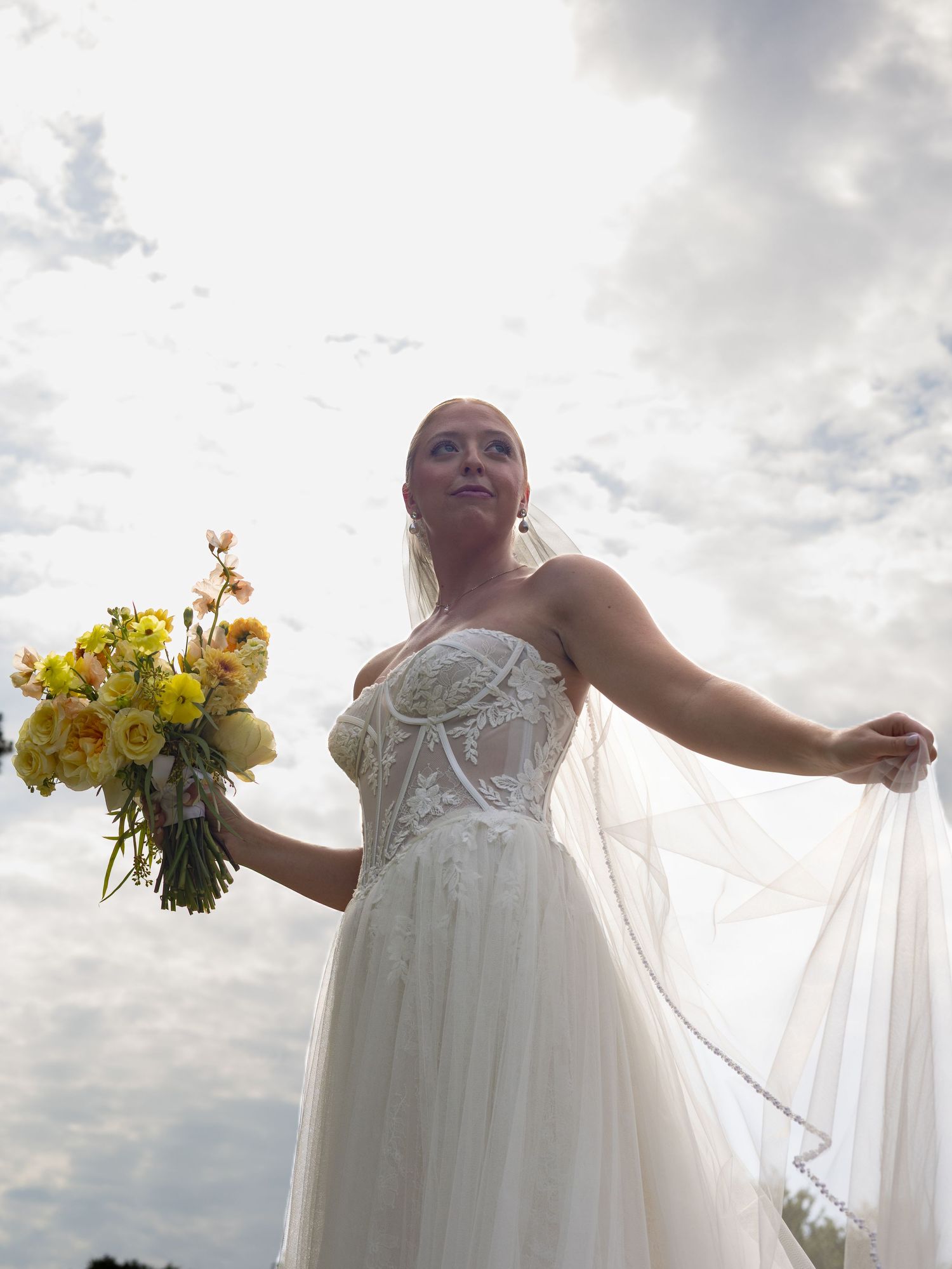 Bride in white lace gown holds yellow bouquet, veil flowing, posed against cloudy sky in elegant outdoor wedding photo.