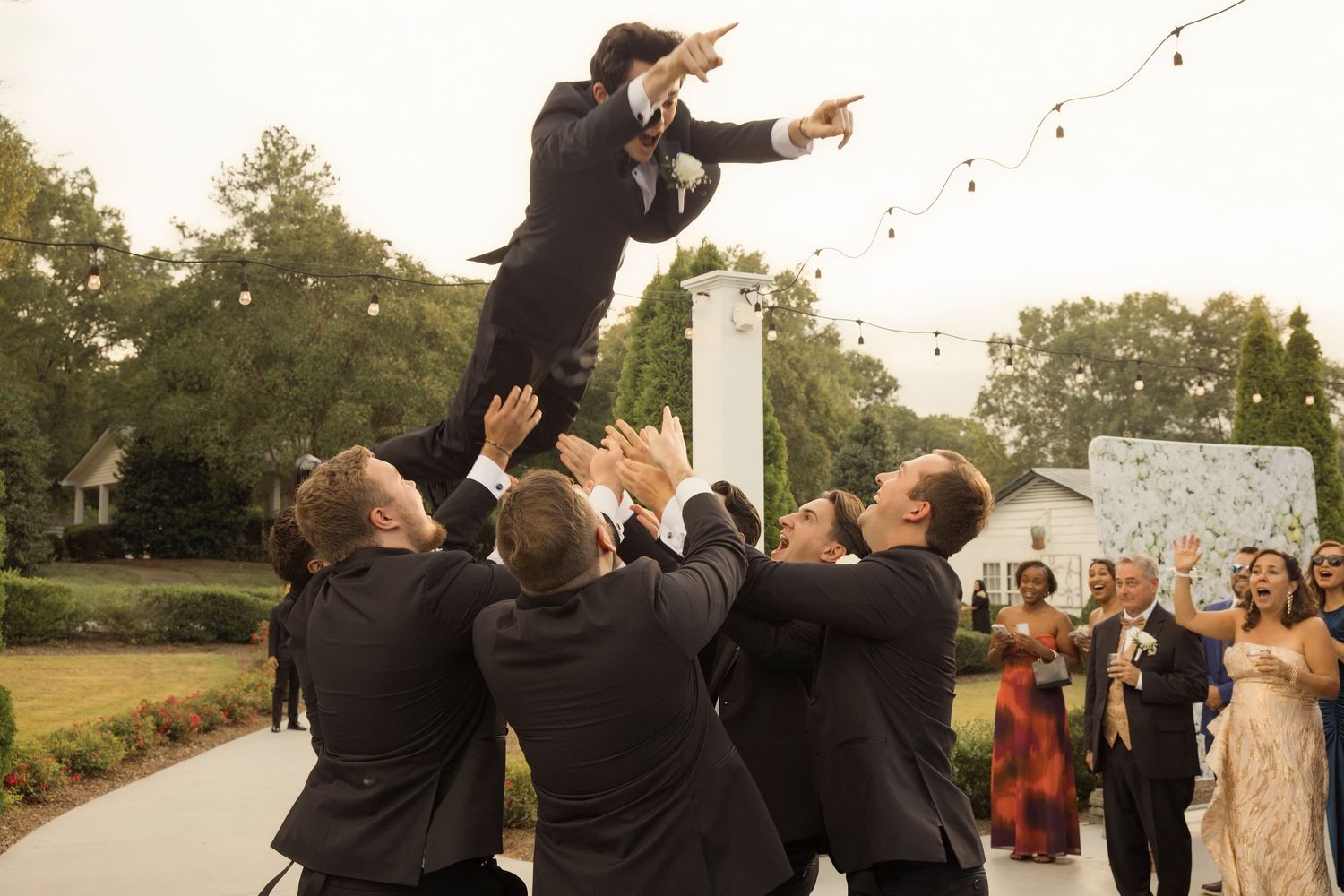 Groomsmen tossing groom in the air during outdoor wedding celebration, guests watching in background.